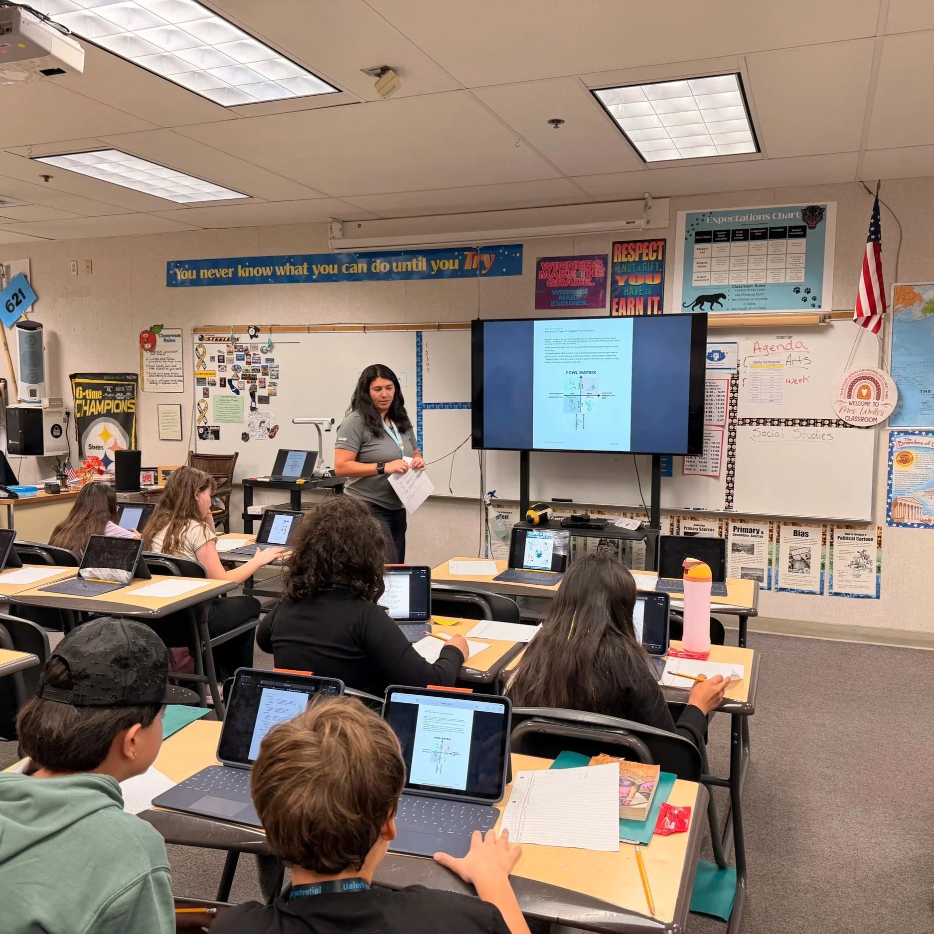 A classroom filled with students working on laptops and notebooks, with a teacher standing at the front near a large screen displaying a presentation on the Fowl Matrix. The classroom has educational posters and an American flag on the wall.