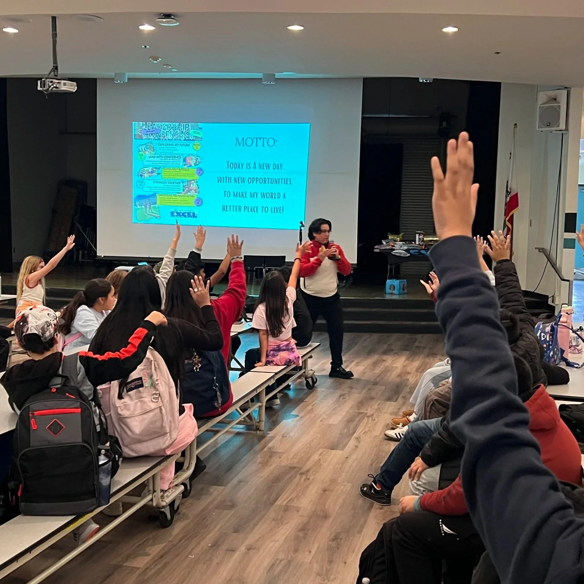 A classroom or conference room with children raising their hands. A presentation on a large screen displays a quote about new opportunities and making the world a better place. Some children are seated on benches, and others are standing, using their phones or talking. The room has a wooden floor and ceiling lights.