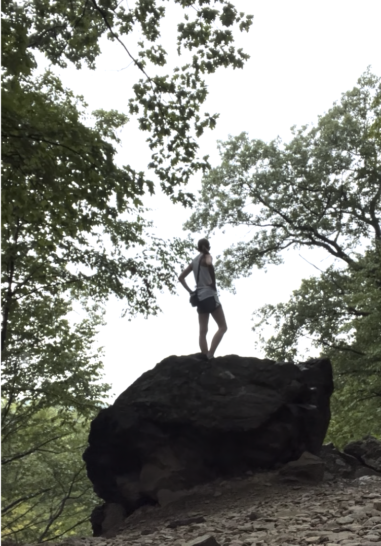Kelly standing on a large rock in the woods looking out over a landscape.