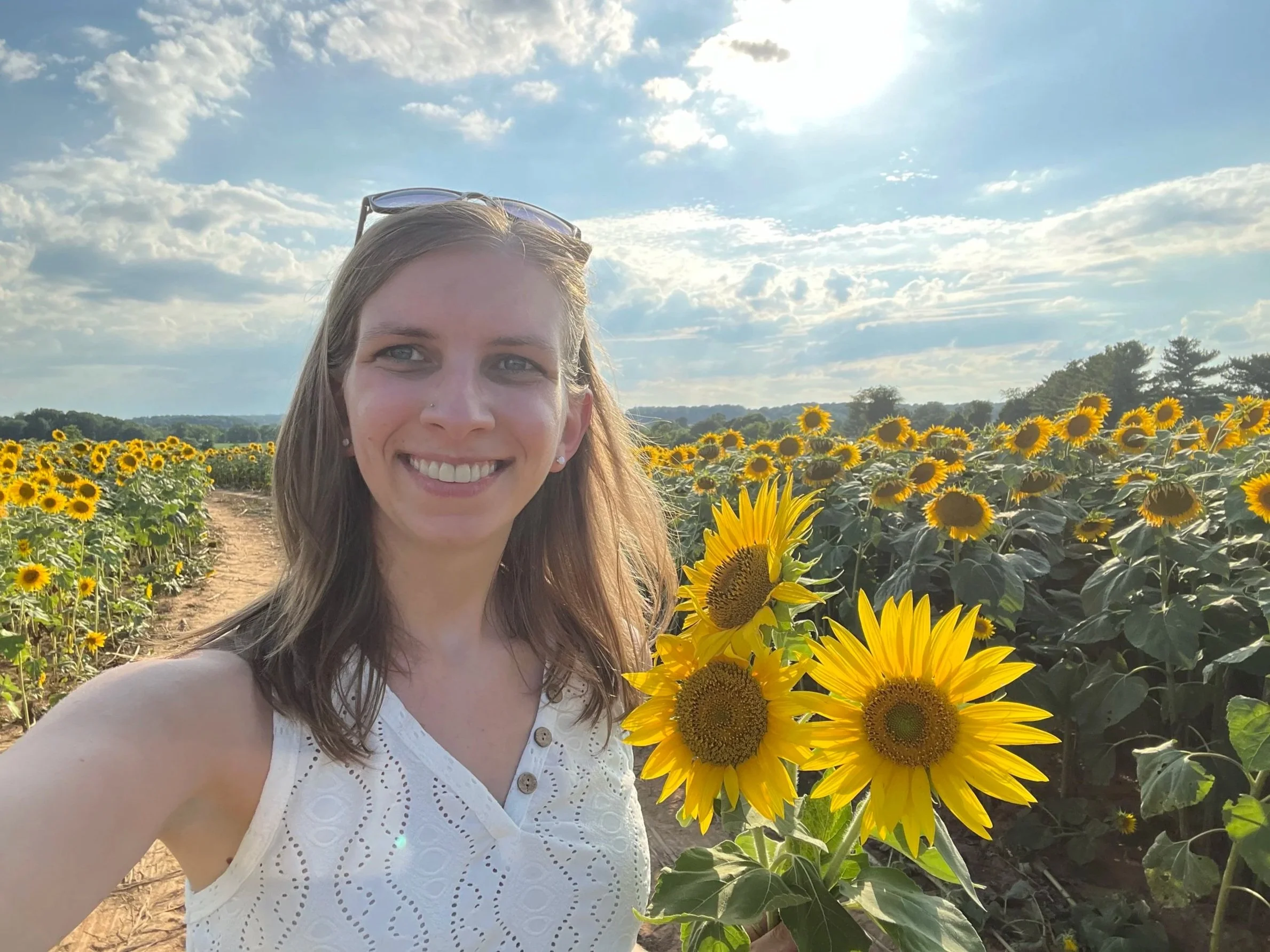 Close up photo of Kelly smiling, holding three sunflowers while standing in a sunny sunflower field.