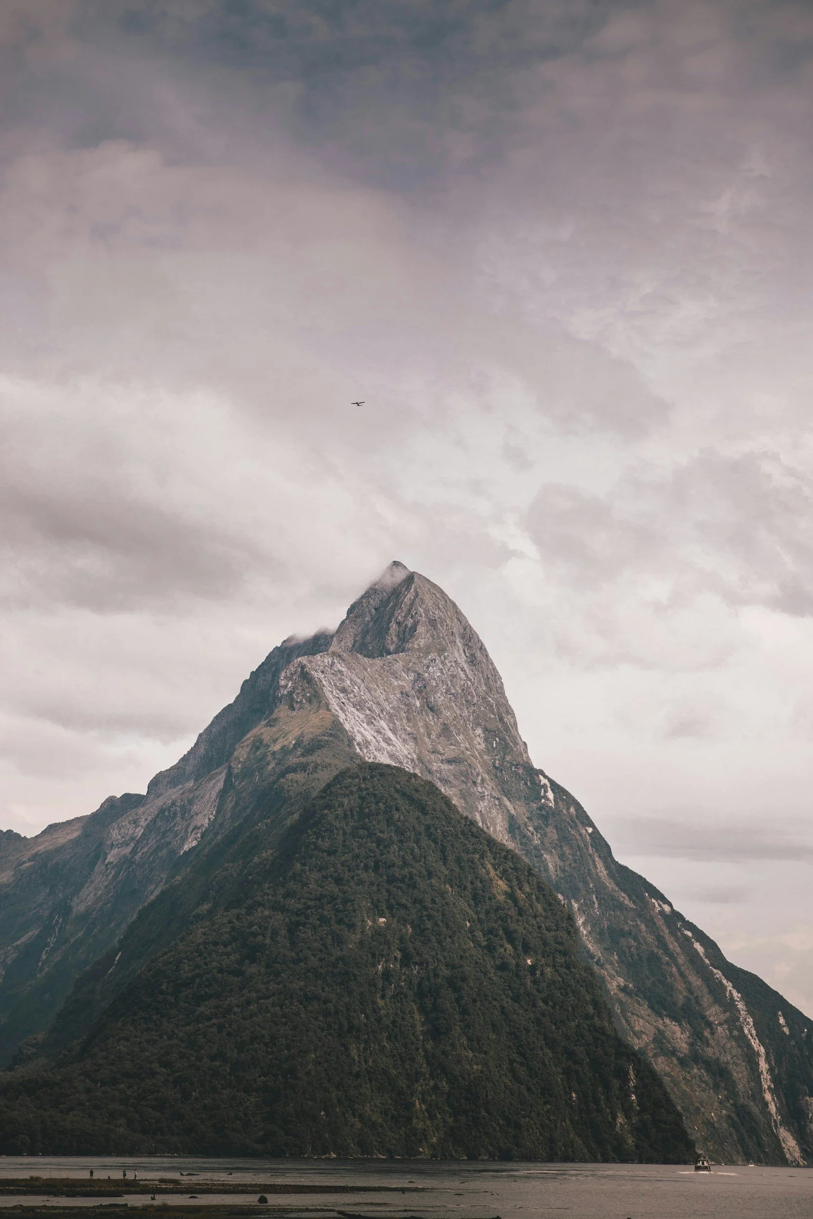 A mountain with a sharp peak surrounded by cloudy sky, with green forested slopes and a body of water with boats at the bottom.