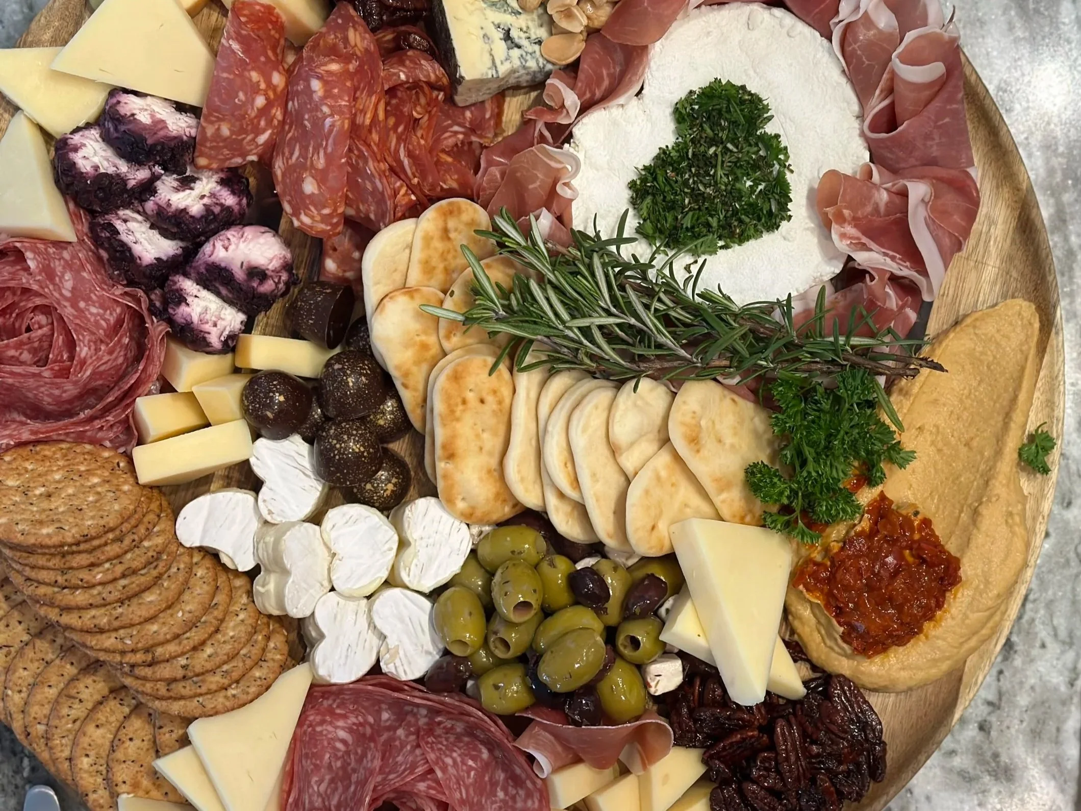 A large round cheese board with assorted sliced cheeses, cured meats, strawberries, grapes, crackers, and garnishing herbs.