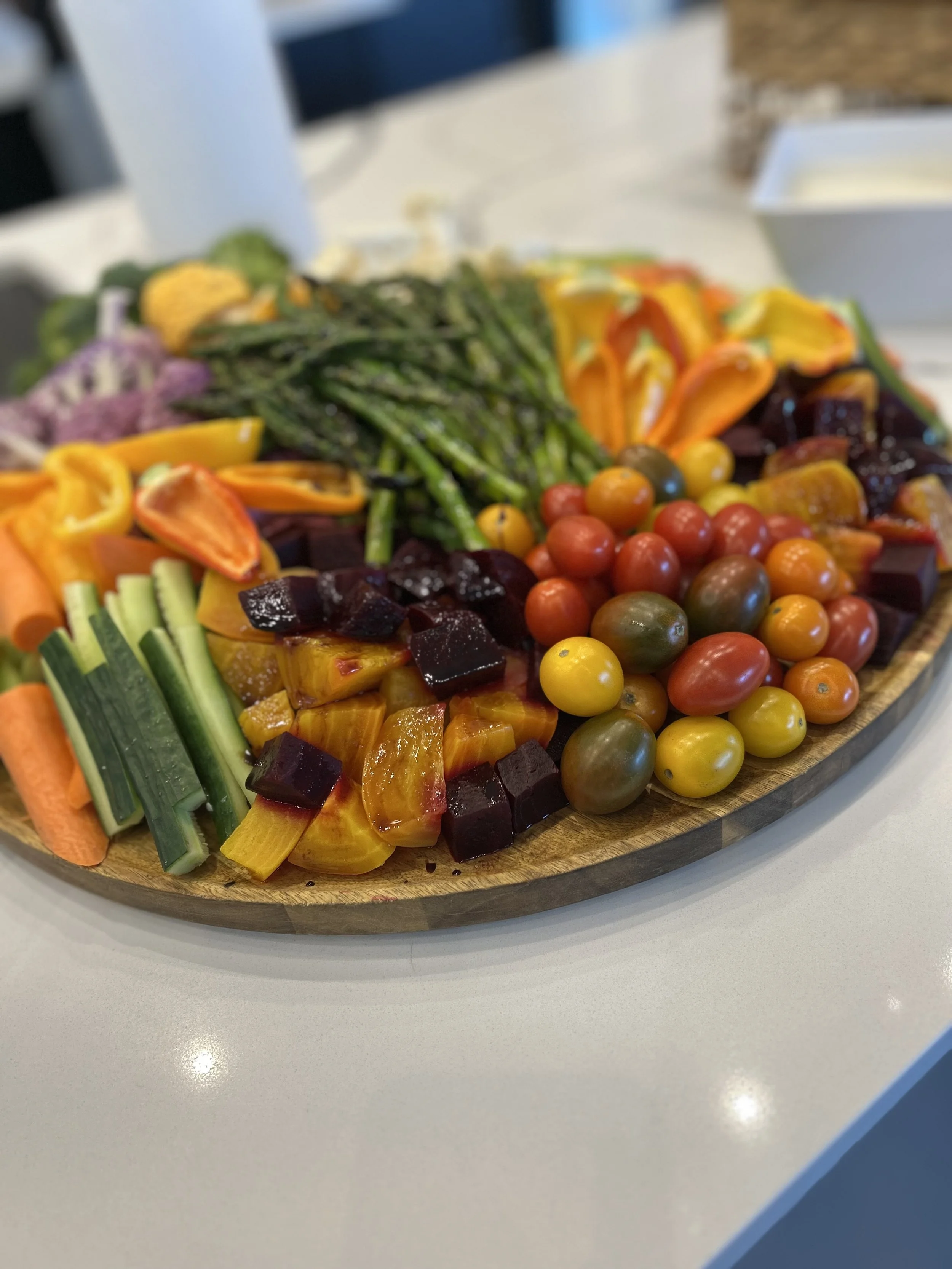 A wooden platter filled with colorful vegetables including cherry tomatoes, sliced carrots, yellow and orange peppers, cucumbers, beets, and asparagus, arranged in sections on a white table.