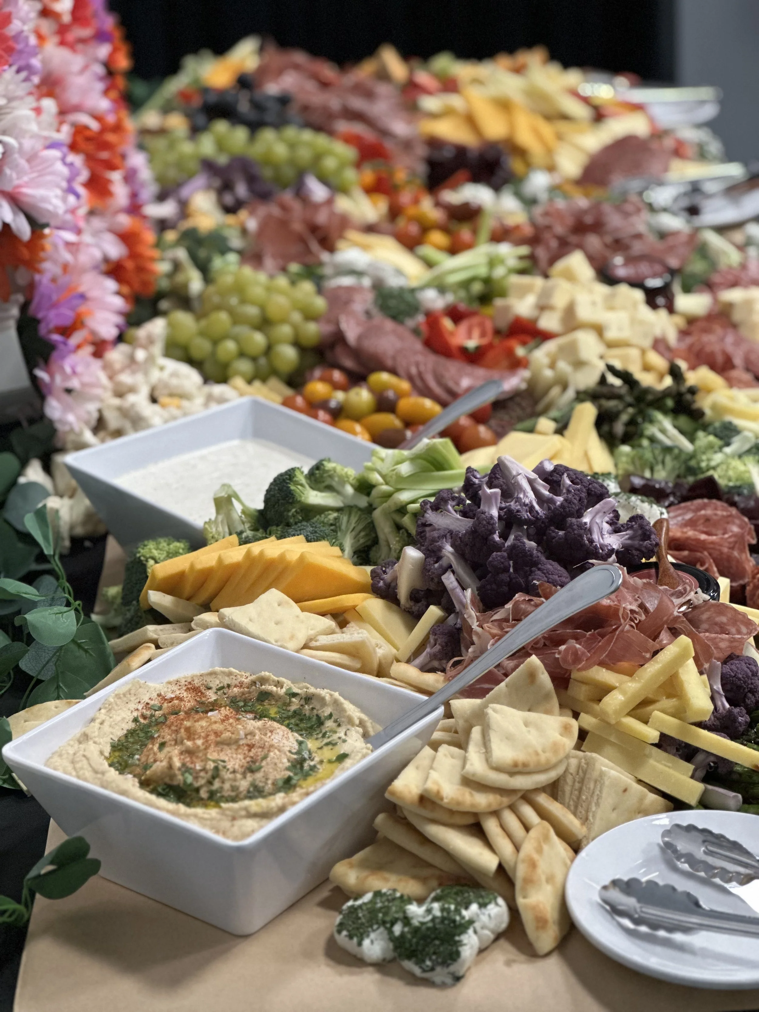 Assorted appetizers, cheeses, grapes, vegetables, hummus, pita bread, and crackers on a buffet table.