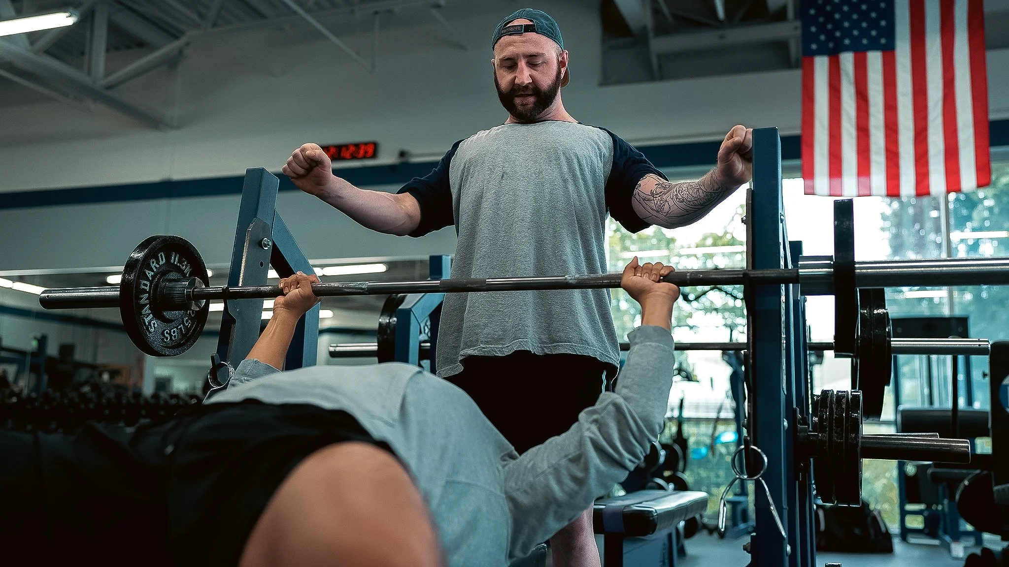 A man with a beard and backwards cap spotting a barbell while lying on a bench press in a gym, with an American flag hanging in the background.
