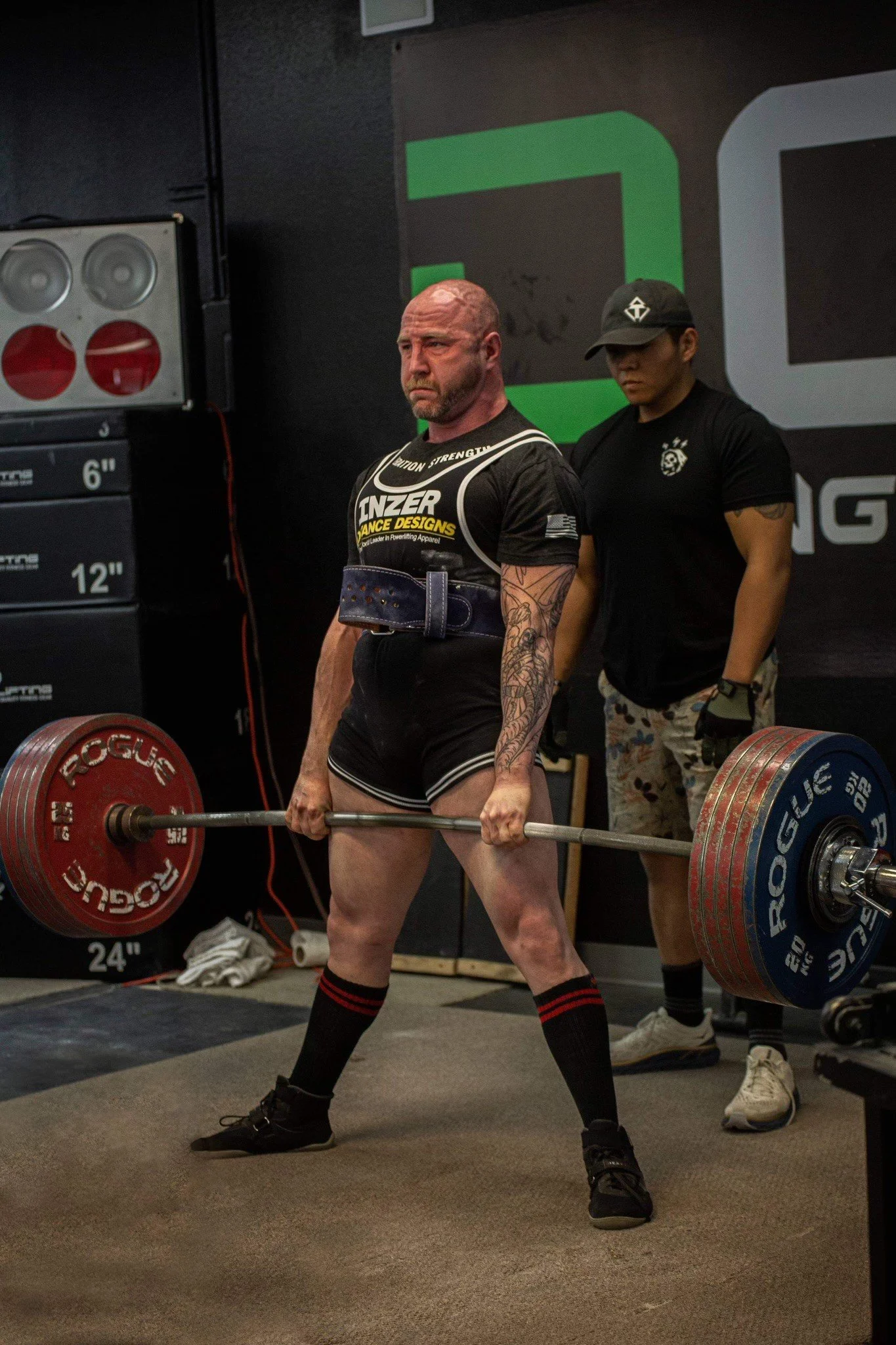 A muscular man with tattoos and a serious expression is lifting a barbell loaded with red and blue weight plates in a gym. Two people stand behind him, one wearing a black t-shirt and camo shorts, and the other in gray athletic shoes. The background includes a large black wall with green and gray geometric designs and gym equipment.