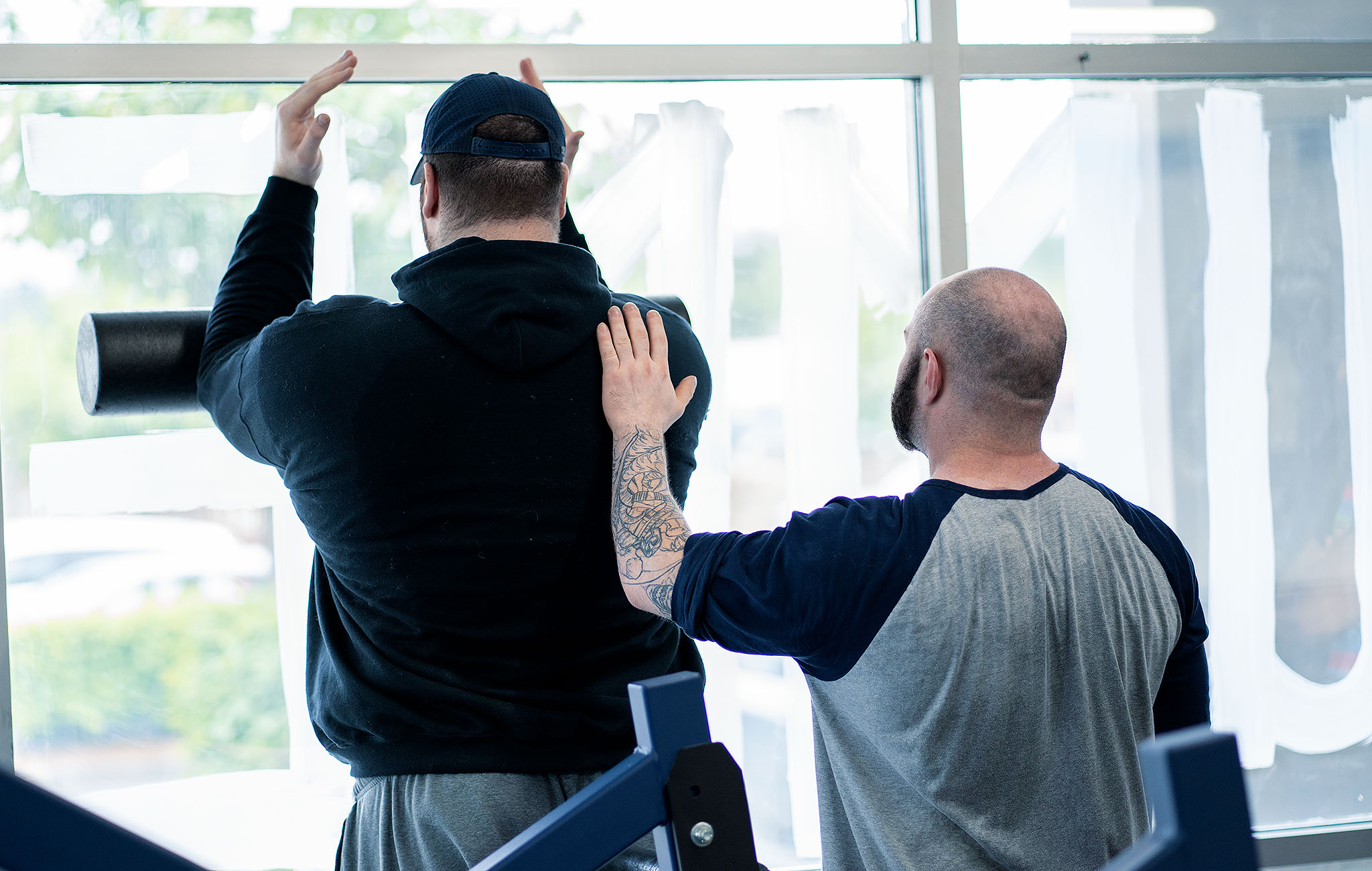 Two men in a physical therapy or gym session, one assisting the other who is stretching near a large window, with exercise equipment visible in the foreground.