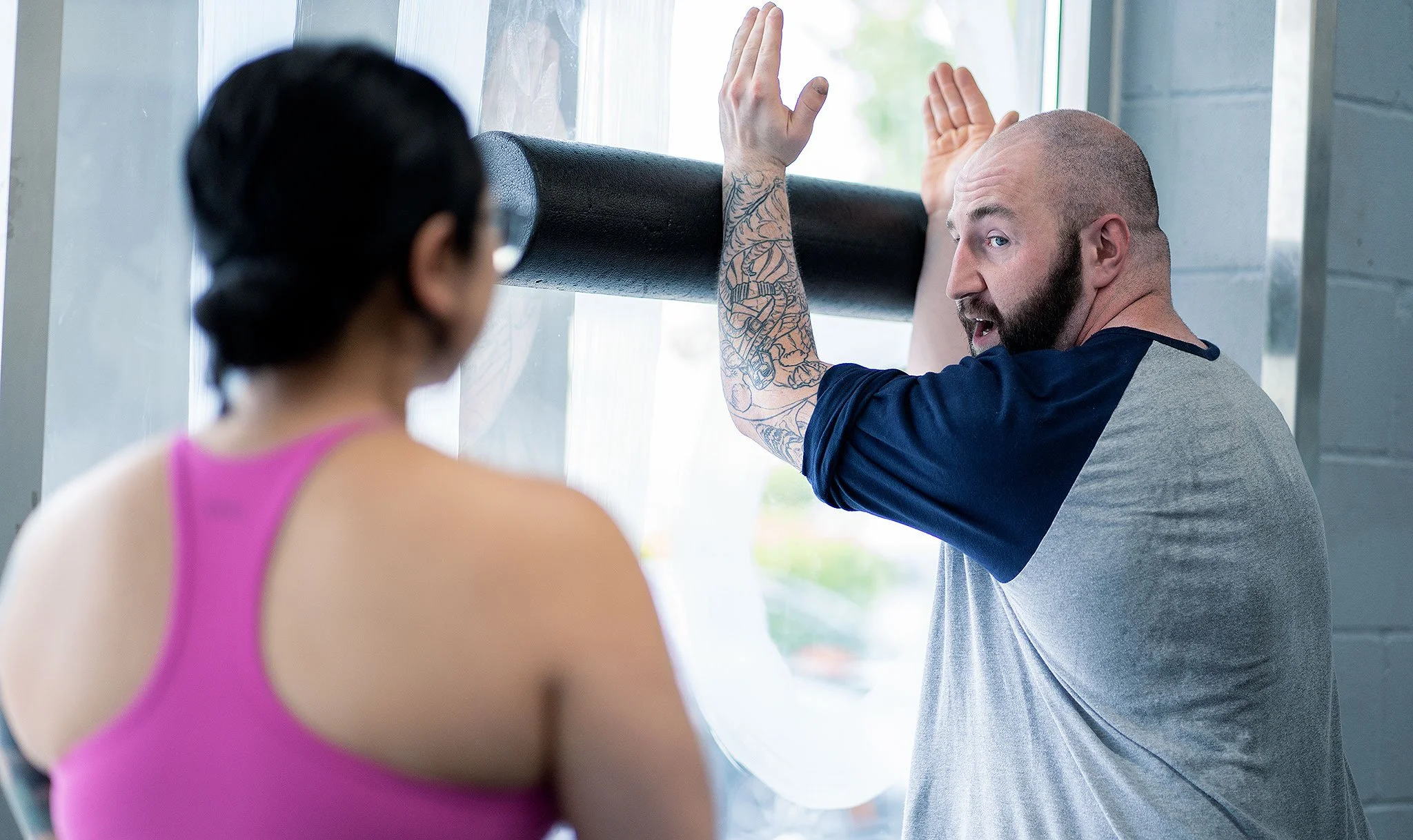 A man with a beard and tattoos on his arm is doing a wall exercise with a woman in a pink top observing. They are indoors near a large window.