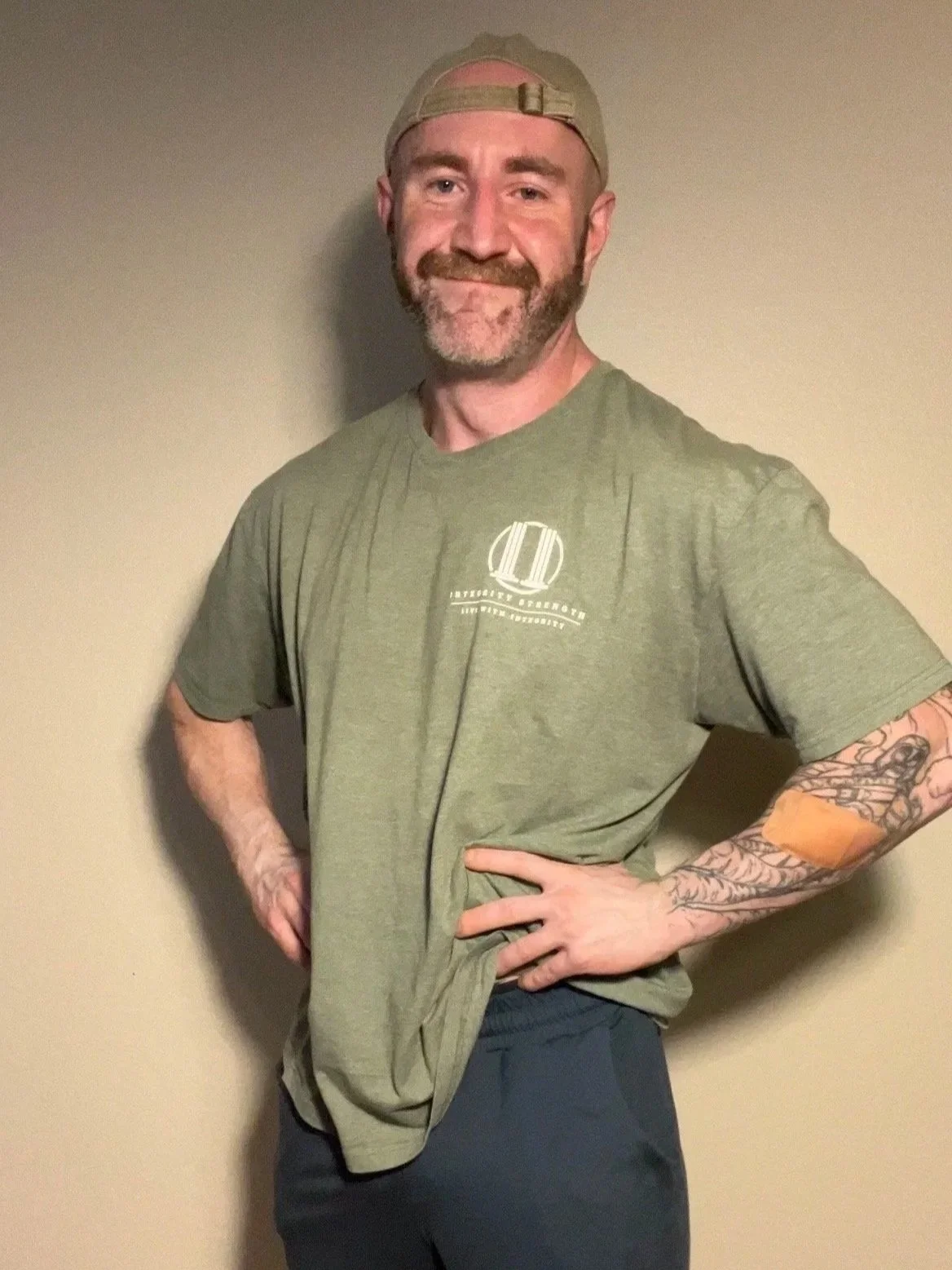 A smiling man with a beard, wearing a khaki cap and a green T-shirt with a logo, standing against a plain wall, with tattoos on his right arm.