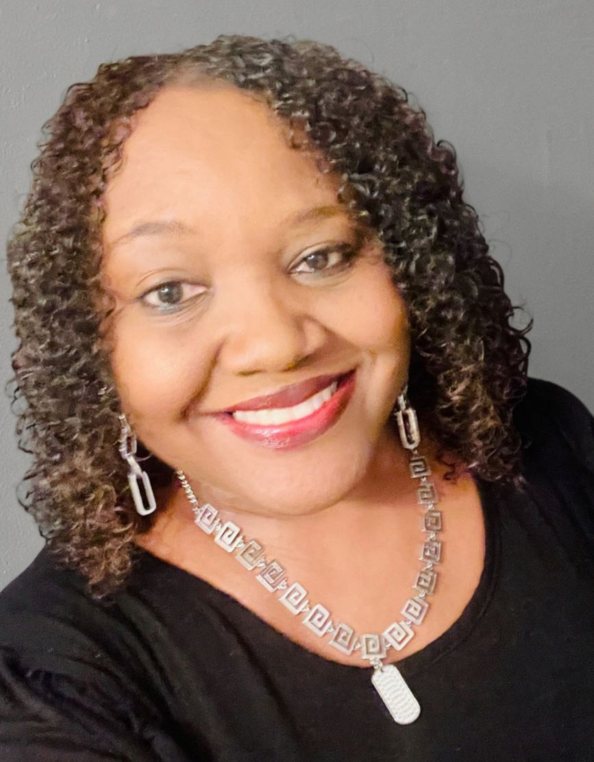 A woman with curly hair smiling, wearing earrings and a necklace with a rectangular pendant, dressed in black against a plain background.