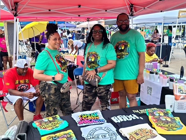 Three people standing behind a table at an outdoor event, wearing matching teal shirts with a colorful design, under a pink canopy. The table displays various colorful T-shirts and merchandise, with more tents and people in the background.
