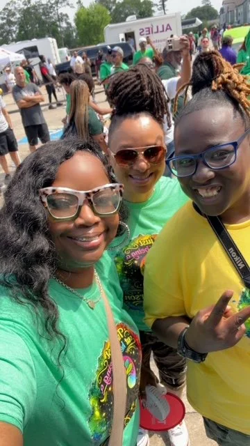 Three women smiling and taking a selfie at an outdoor event with many people, tents, and buses in the background.