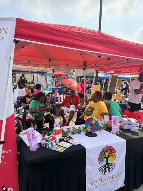 People sitting at a booth under a red canopy tent at an outdoor event, with promotional items and a banner for Whole Person Wellness Collective.