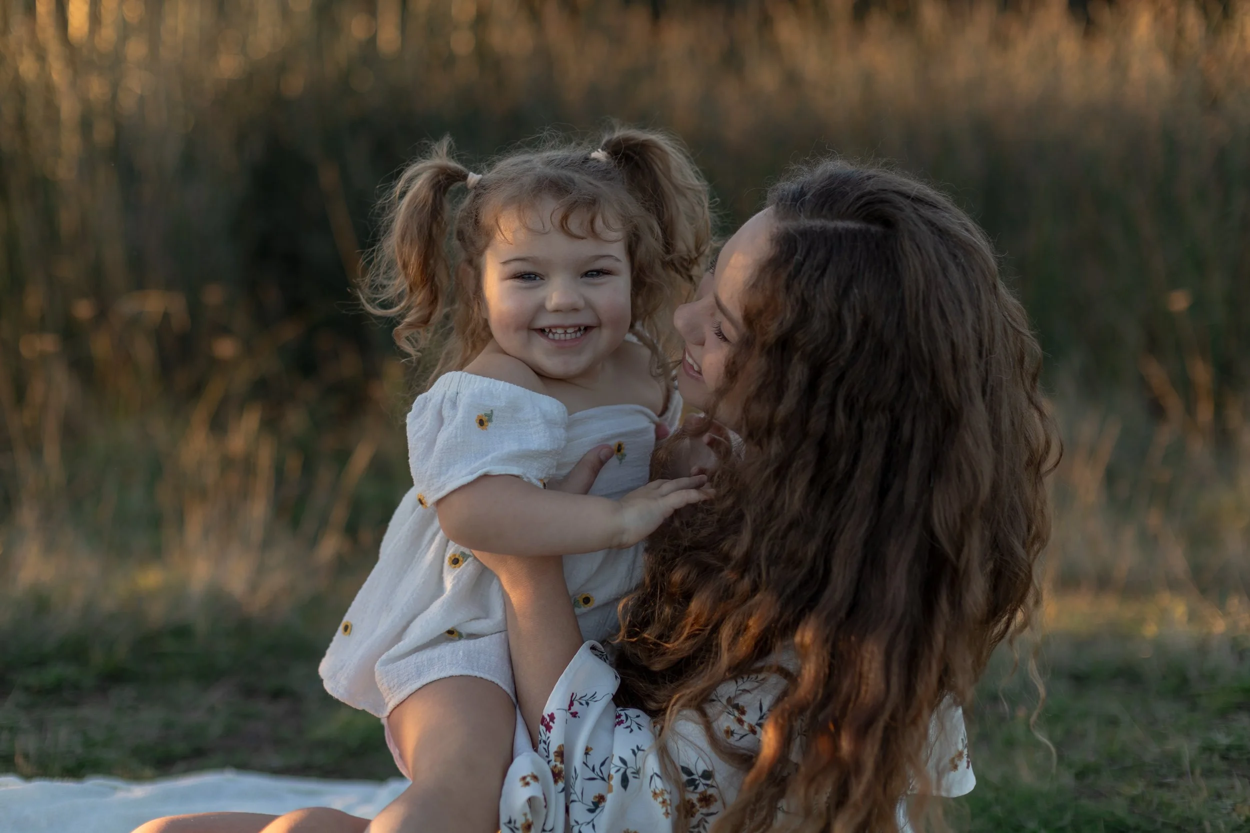 A woman holding a smiling young girl outdoors during sunset, with tall grass in the background.