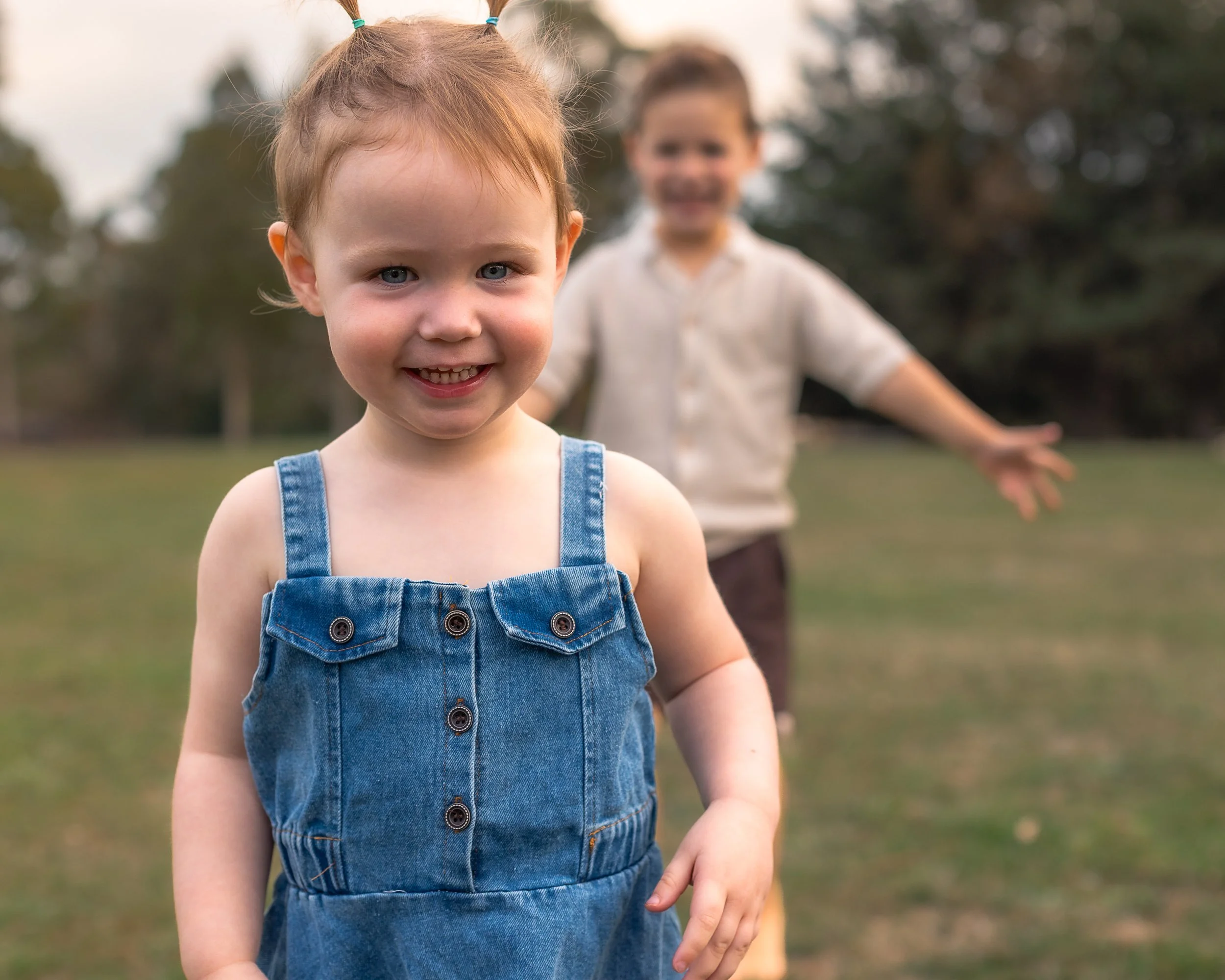 Close-up of a young girl with blue eyes and red hair in pigtails, smiling, wearing a denim dress with buttons. In the background, a boy with brown hair, also smiling, wearing a light-colored shirt, is out of focus outdoors in a park or garden setting