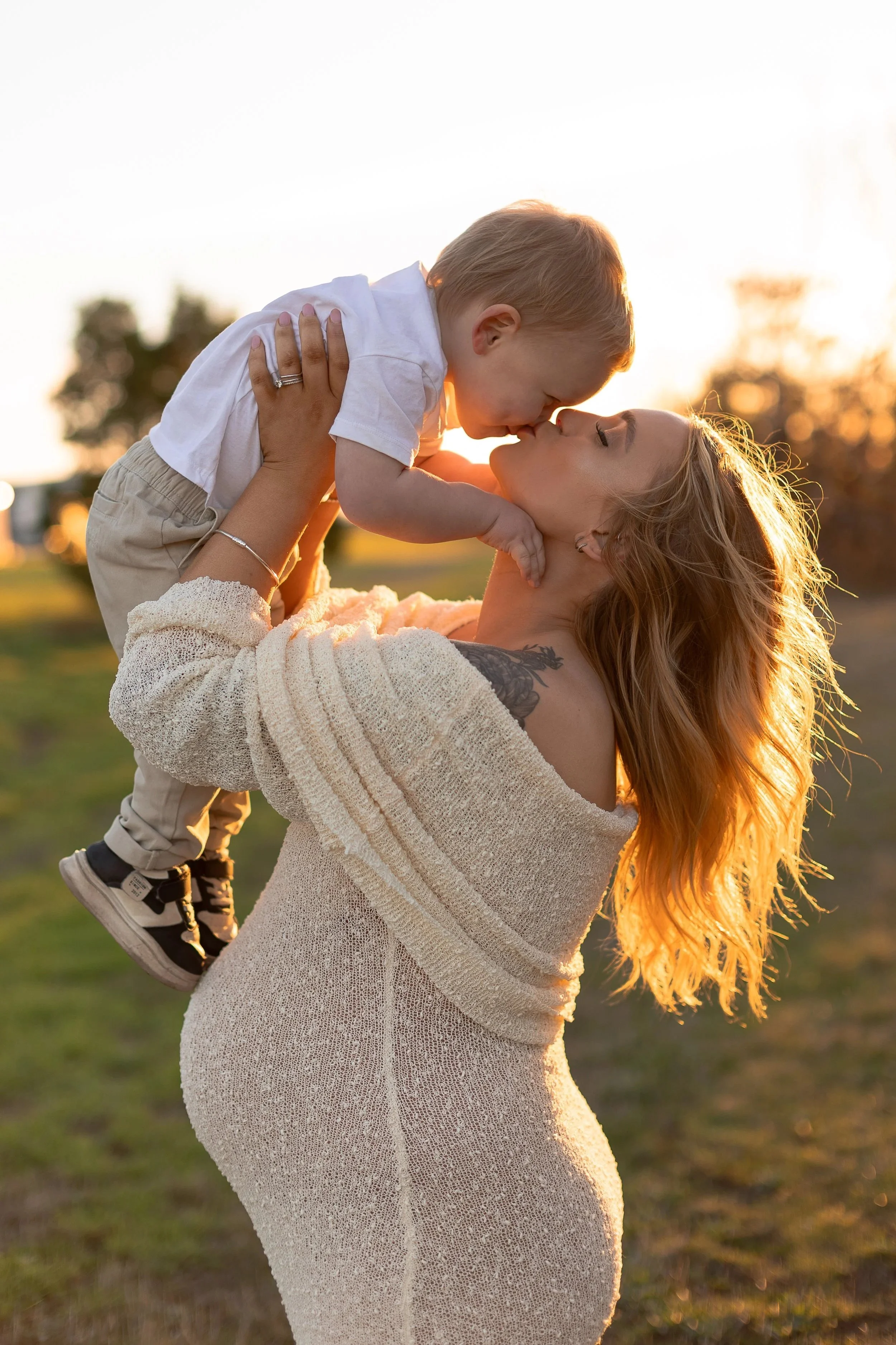 A woman with long red hair holding a young boy up as they lean in for a kiss outdoors during sunset.