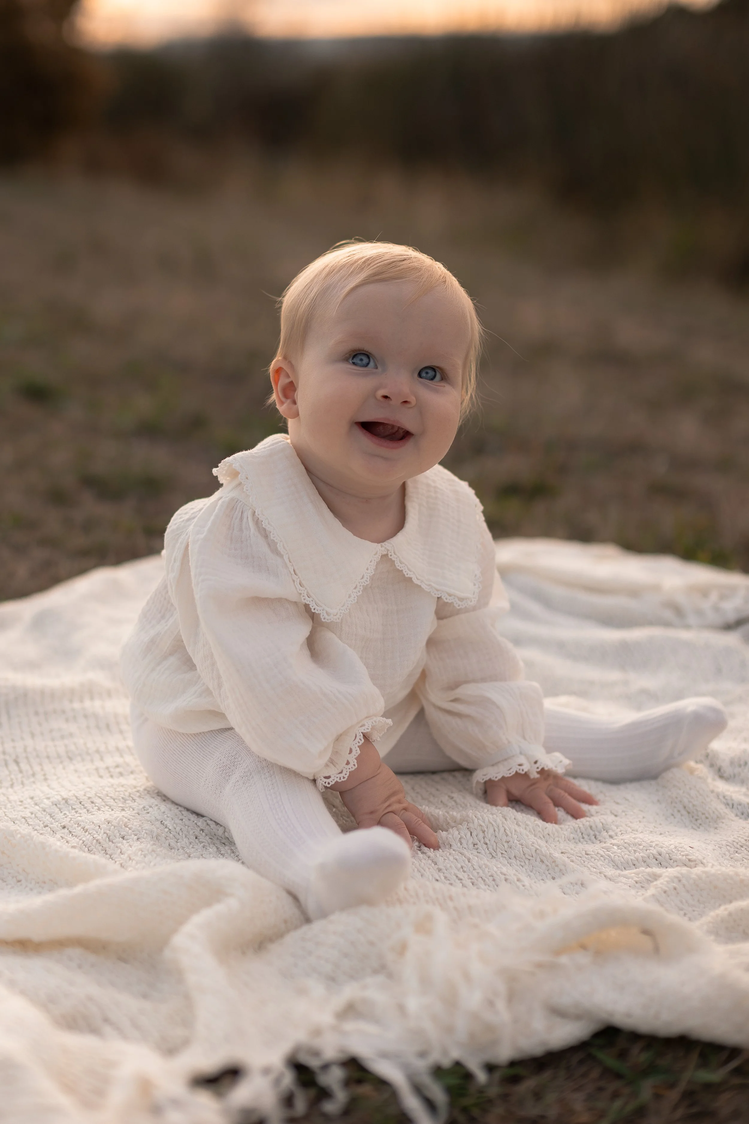 A smiling baby with blonde hair and blue eyes, wearing a cream-colored outfit with lace details, sitting on a white blanket outdoors during sunset, with a blurred natural background.