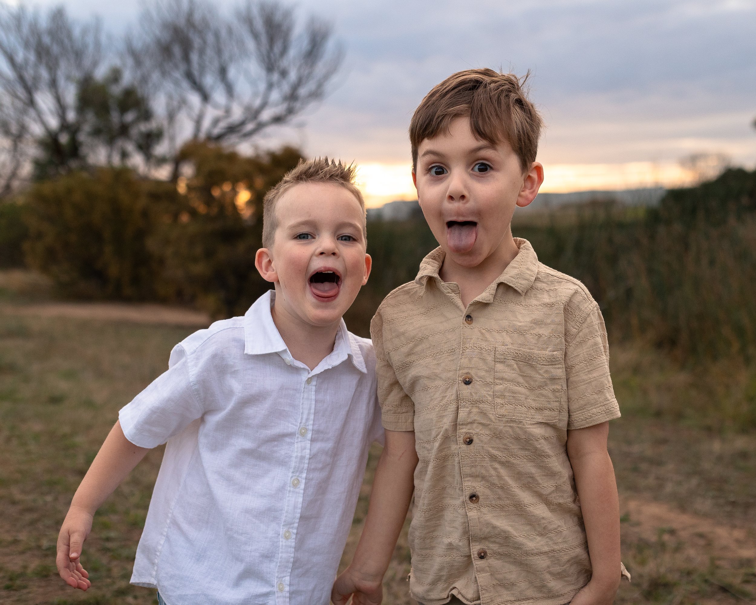 Two young boys are standing outdoors at sunset, making silly faces. One boy is sticking out his tongue, and the other has his mouth open wide, with a surprised expression. They are dressed casually, with one in a white shirt and the other in a beige 