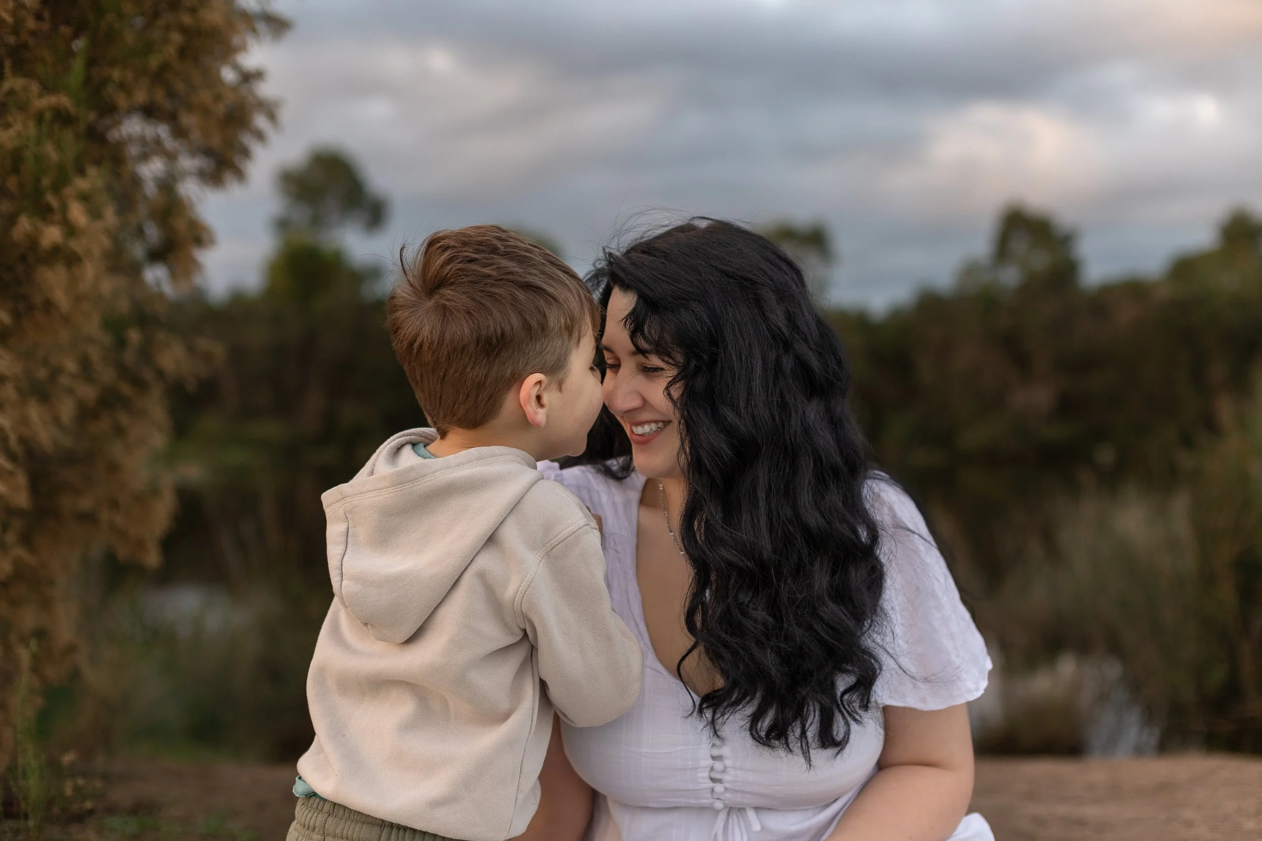 A woman with long black hair smiling as she holds a young boy with short brown hair close to her face outdoors. They appear to be sharing a tender moment, with the background showing trees and a cloudy sky.