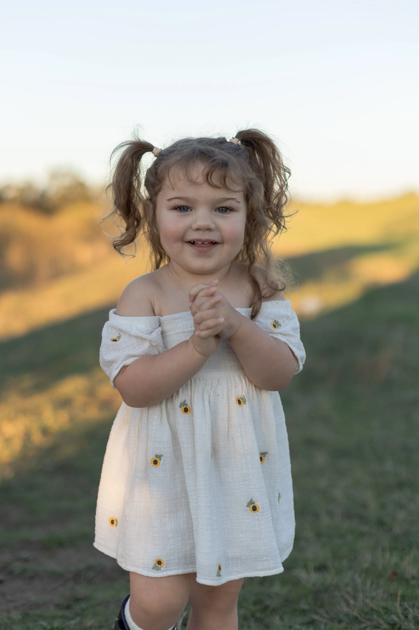 A young girl with curly hair tied in pigtails, wearing a white dress with sunflower embroidery, standing outdoors on grass, smiling with hands clasped.