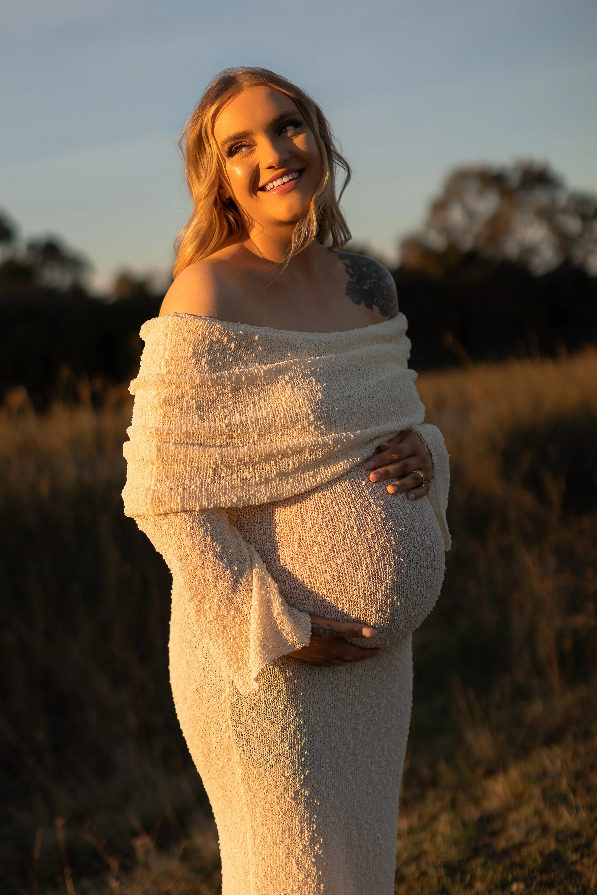 A pregnant woman standing outdoors at sunset, wearing a textured off-shoulder beige dress, smiling and looking to the side with her hair styled in loose waves and a tattoo of flowers visible on her shoulder.