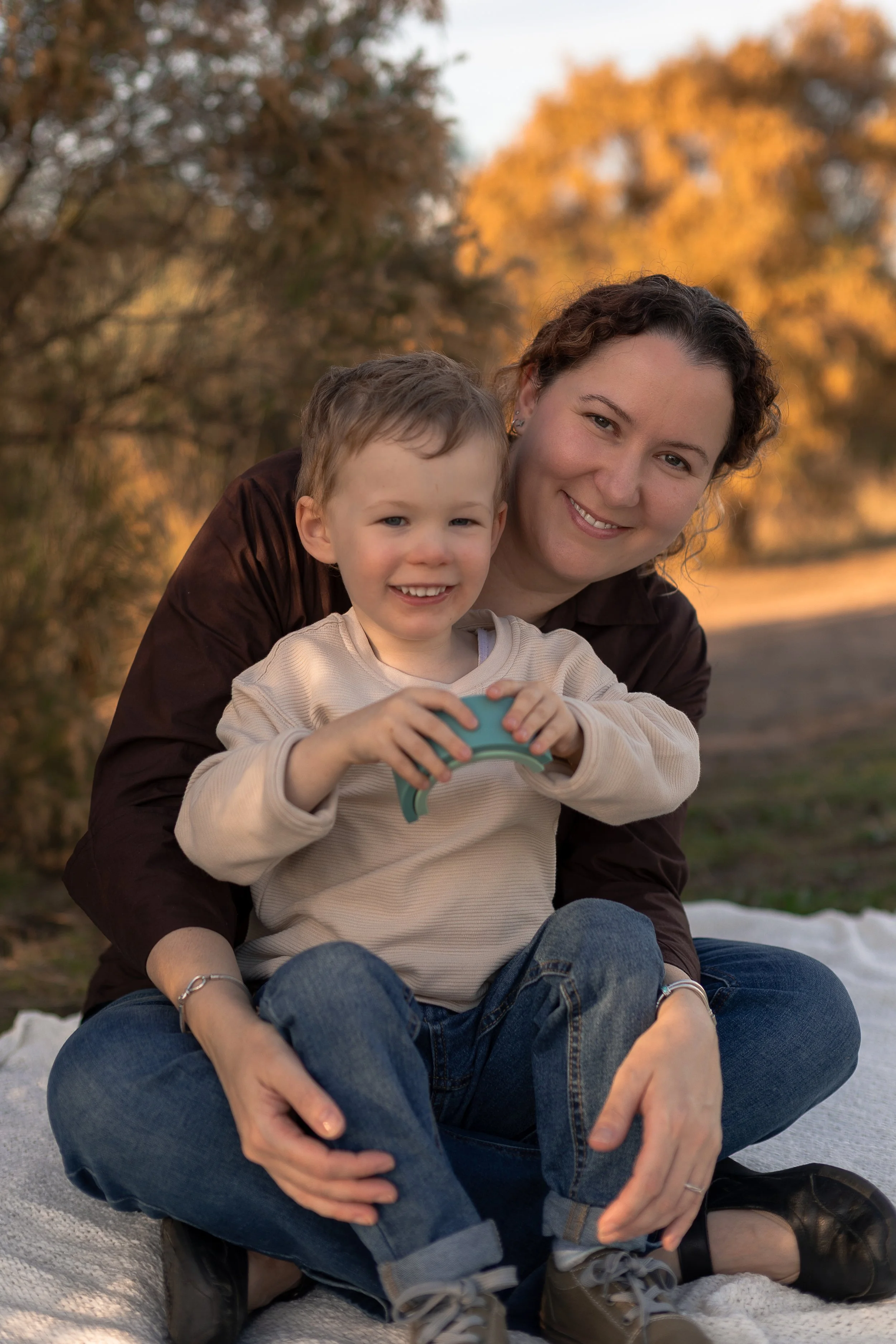 A smiling woman and a boy sitting on a blanket outdoors during autumn, with trees and orange foliage in the background.