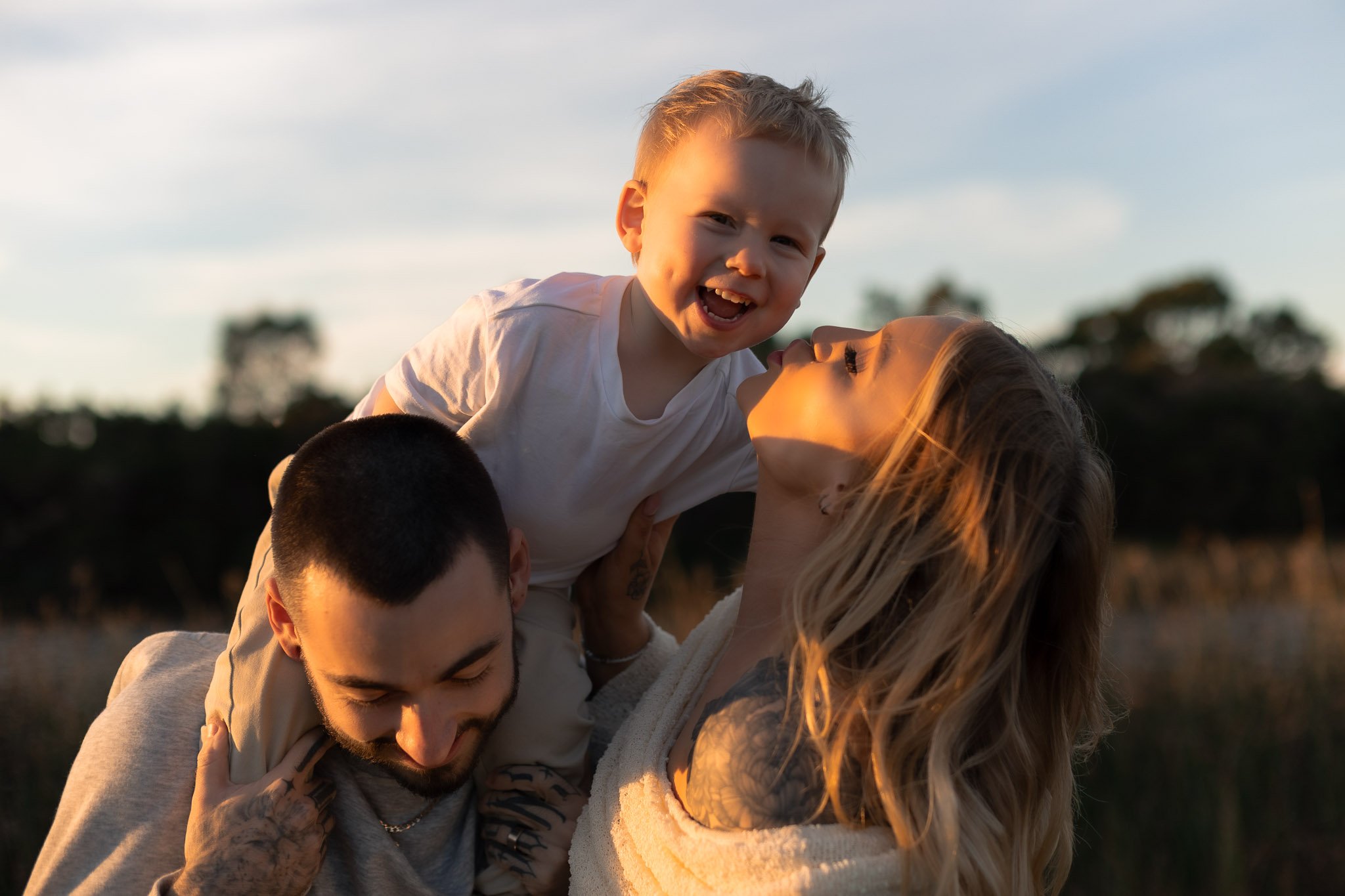 Group of four young people outdoors, with one lifting a smiling child in the air at sunset, blurred trees in the background.