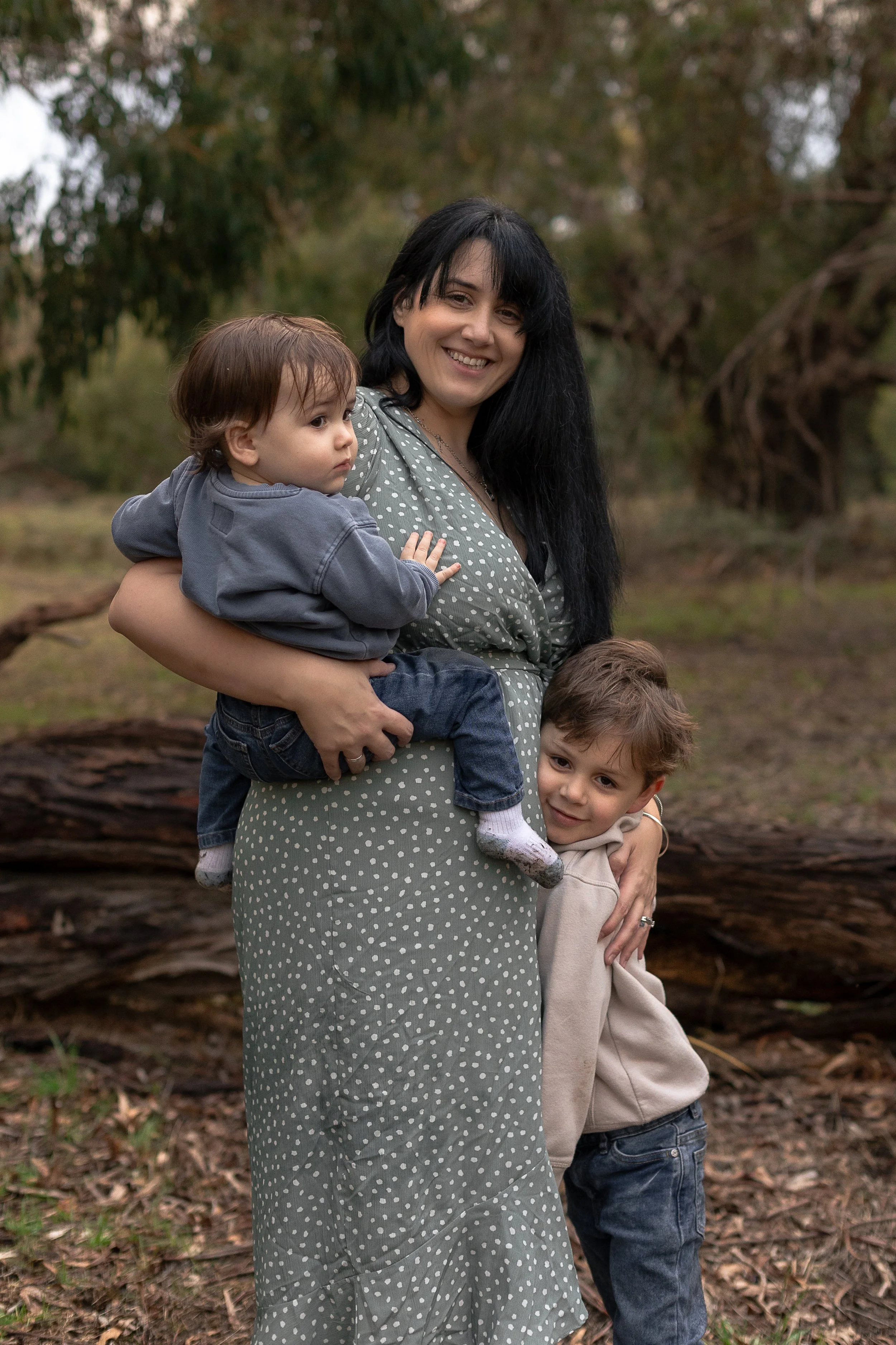 A smiling woman wearing a gray polka dot dress holds a young boy with brown hair and a gray hoodie on her lap in a wooded outdoor setting with trees and fallen logs.