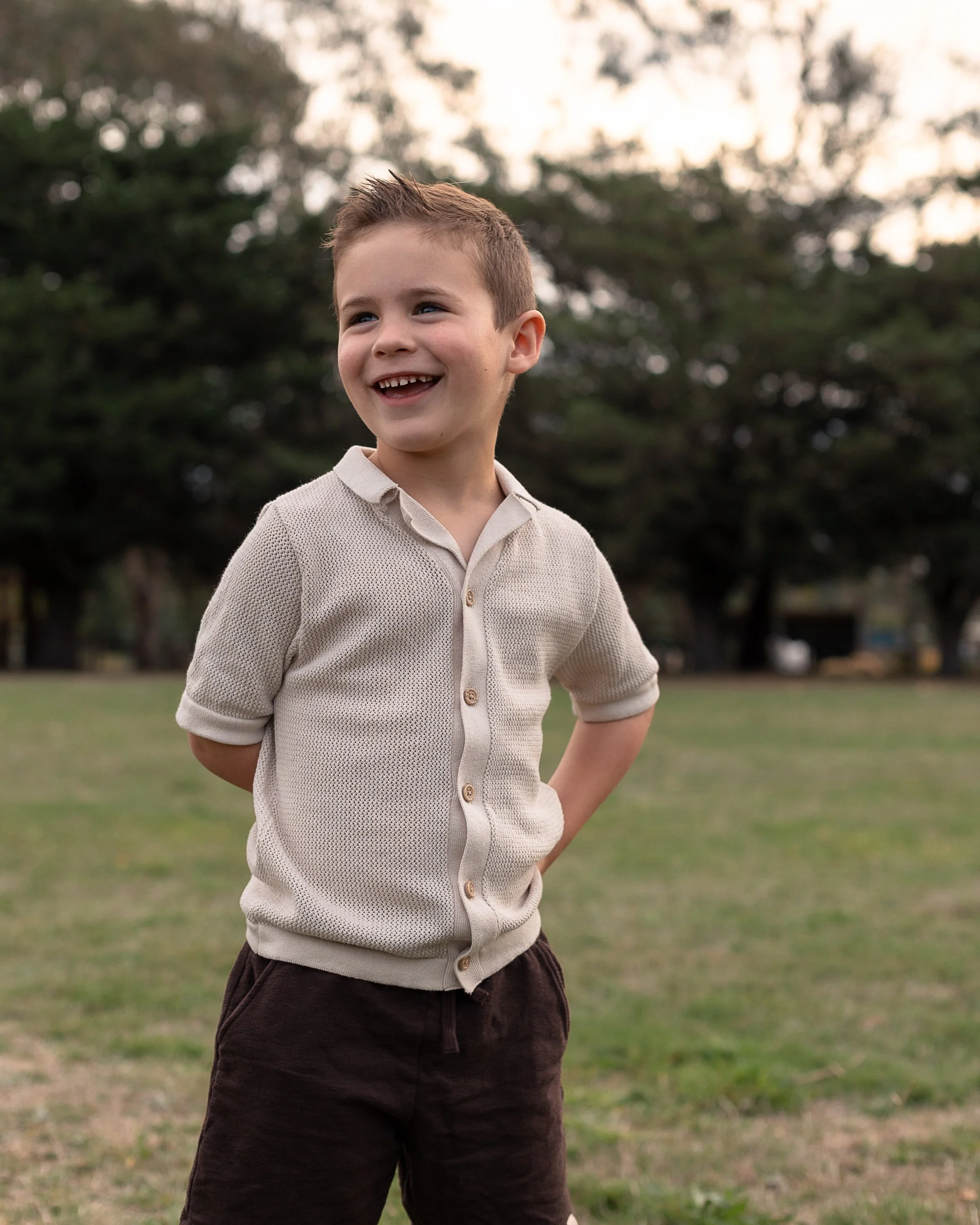 A young boy with short light brown hair and a big smile stands outside in a grassy area with trees in the background, wearing a beige sweater and dark pants.