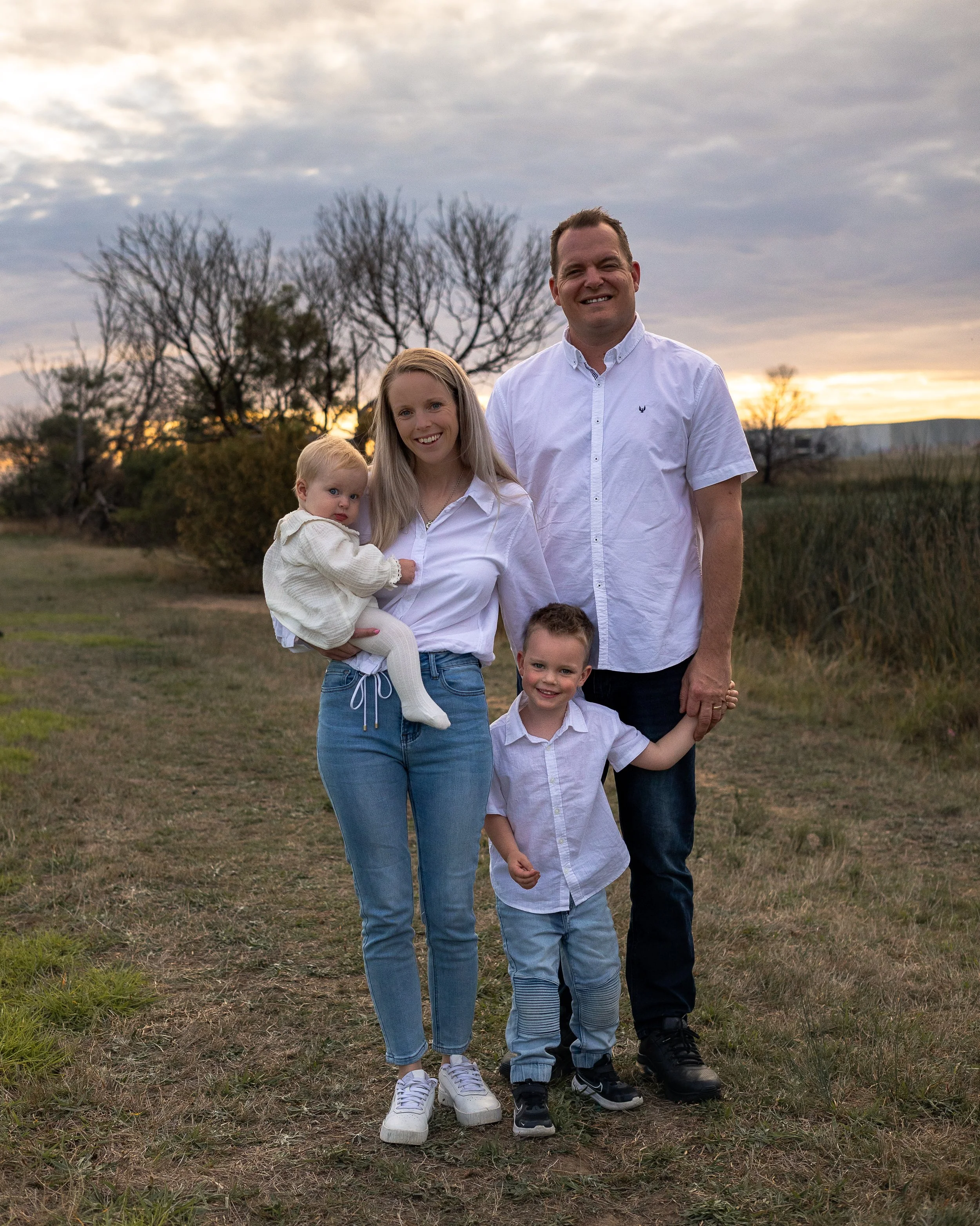 A family of five outdoor photo during sunset, consisting of a woman, a man, and three children, standing on grass with trees and buildings in the background.