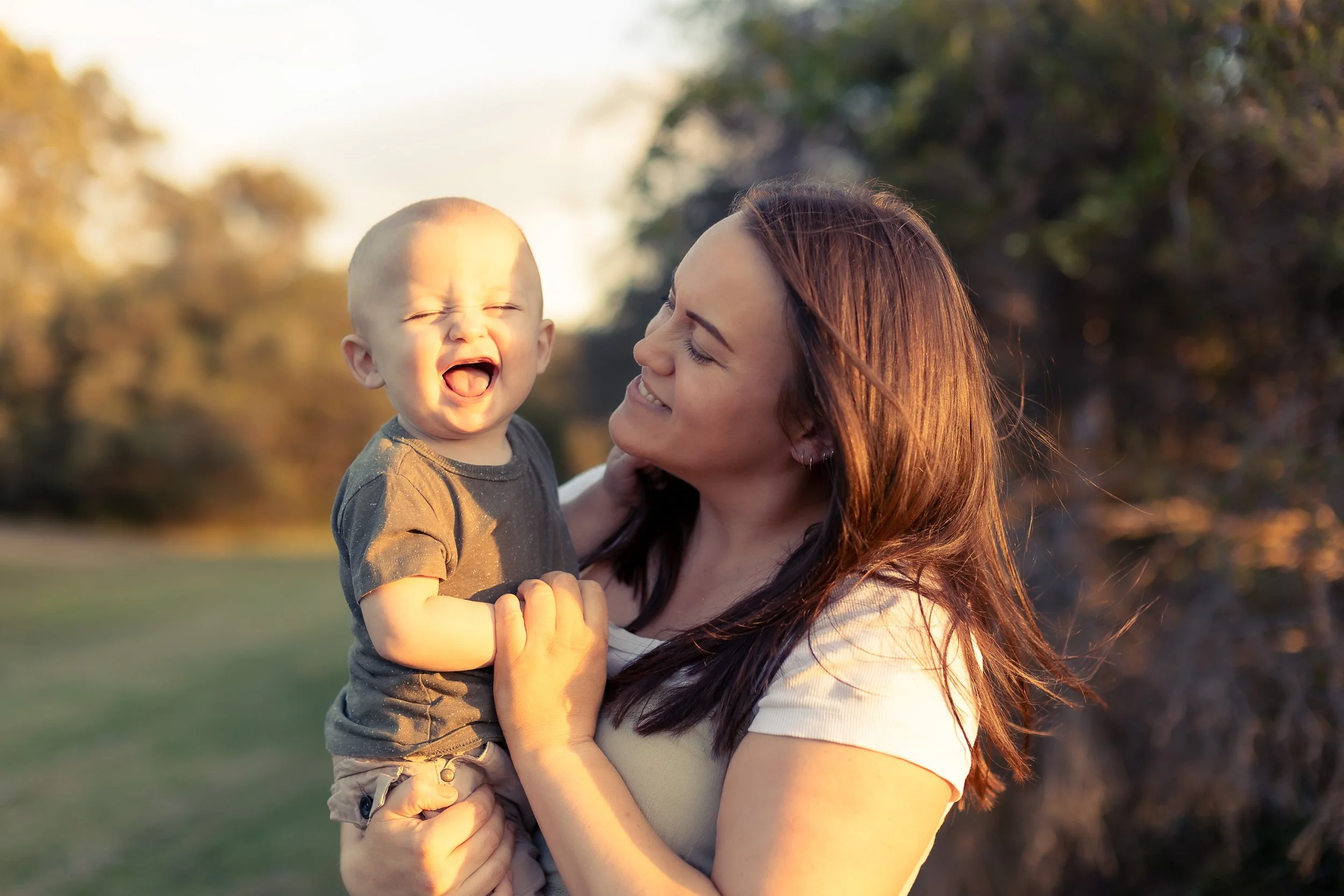A woman holding a happy young boy outdoors during sunset with trees in the background.
