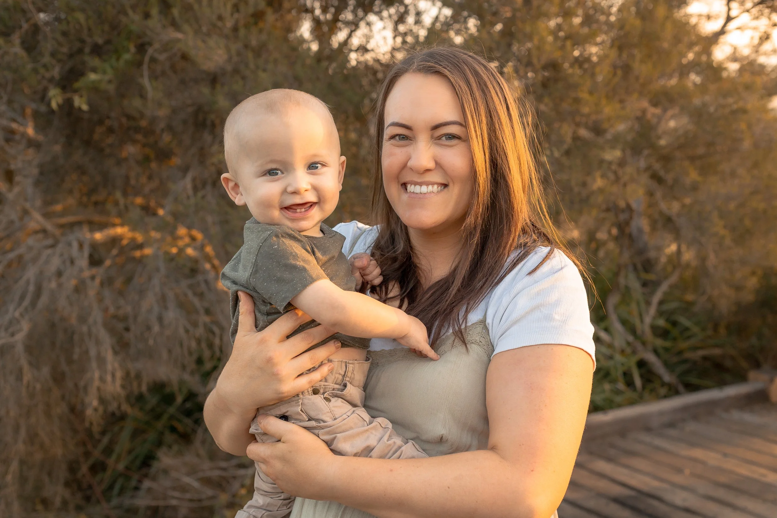 A woman holding a smiling toddler outdoors during sunset, with trees in the background.