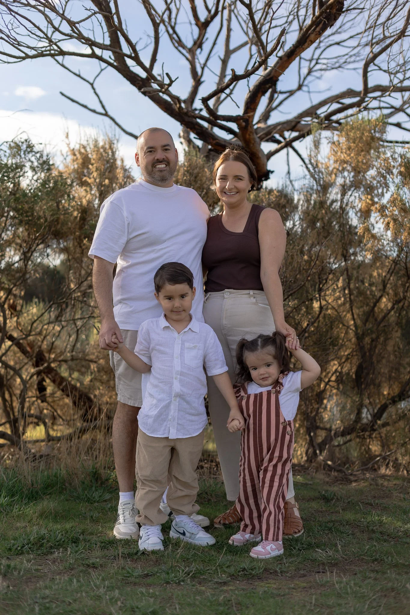 A family of five standing outdoors in front of a leafless tree in a park, with a man, woman, and three children smiling and holding hands.
