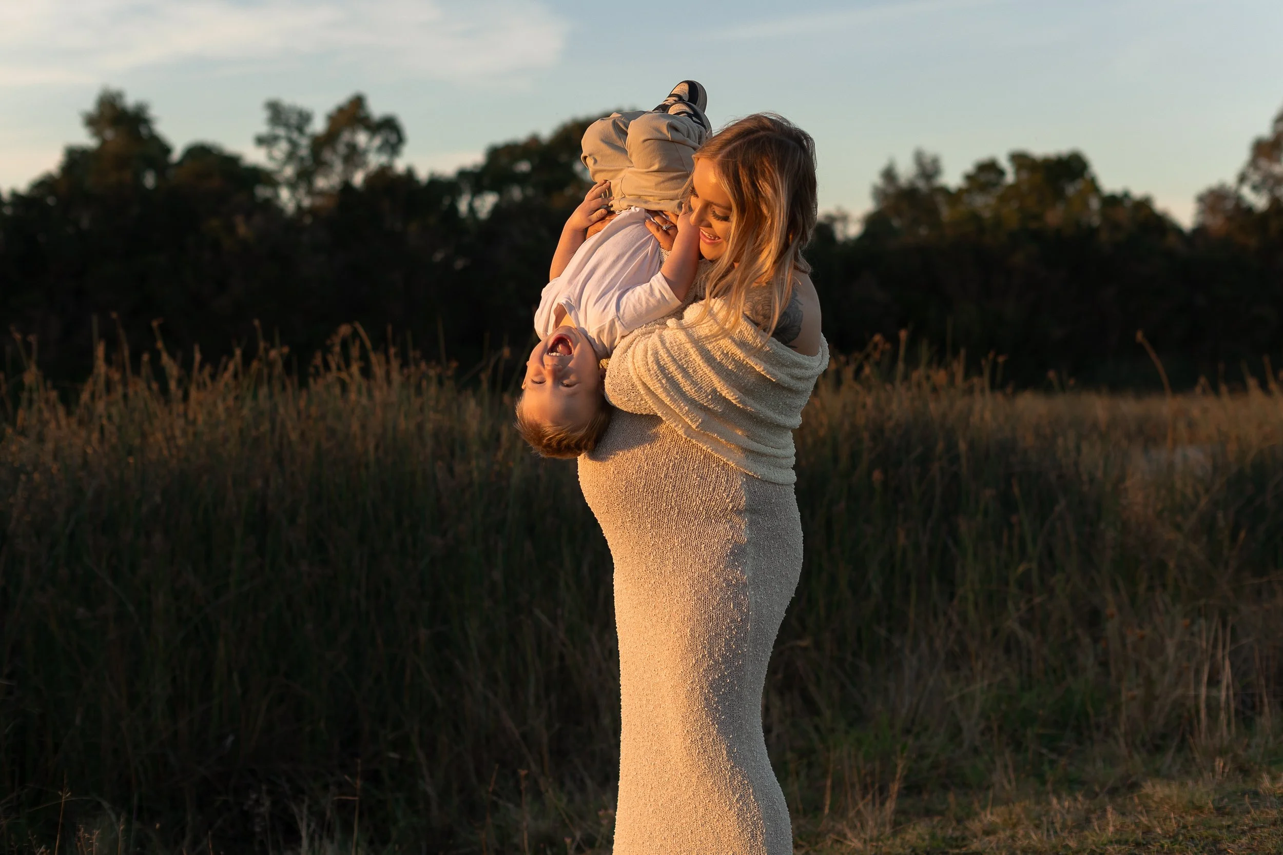 A woman holding a laughing young boy upside down in a field during sunset.