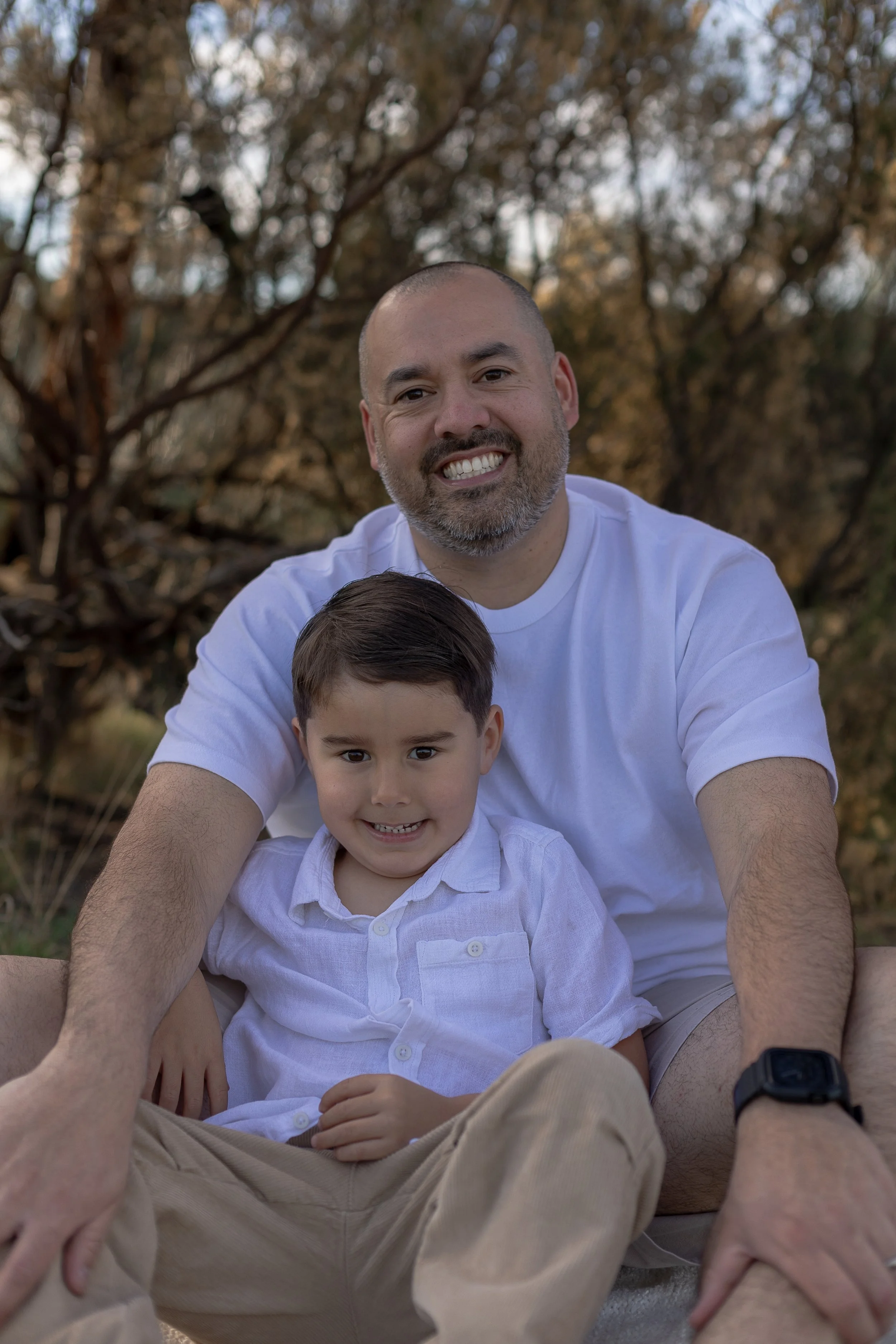 A man and a young boy sitting outdoors in front of trees, smiling for a photo. The man has a beard and is wearing a white t-shirt, while the boy has short hair and is wearing a white button-up shirt.