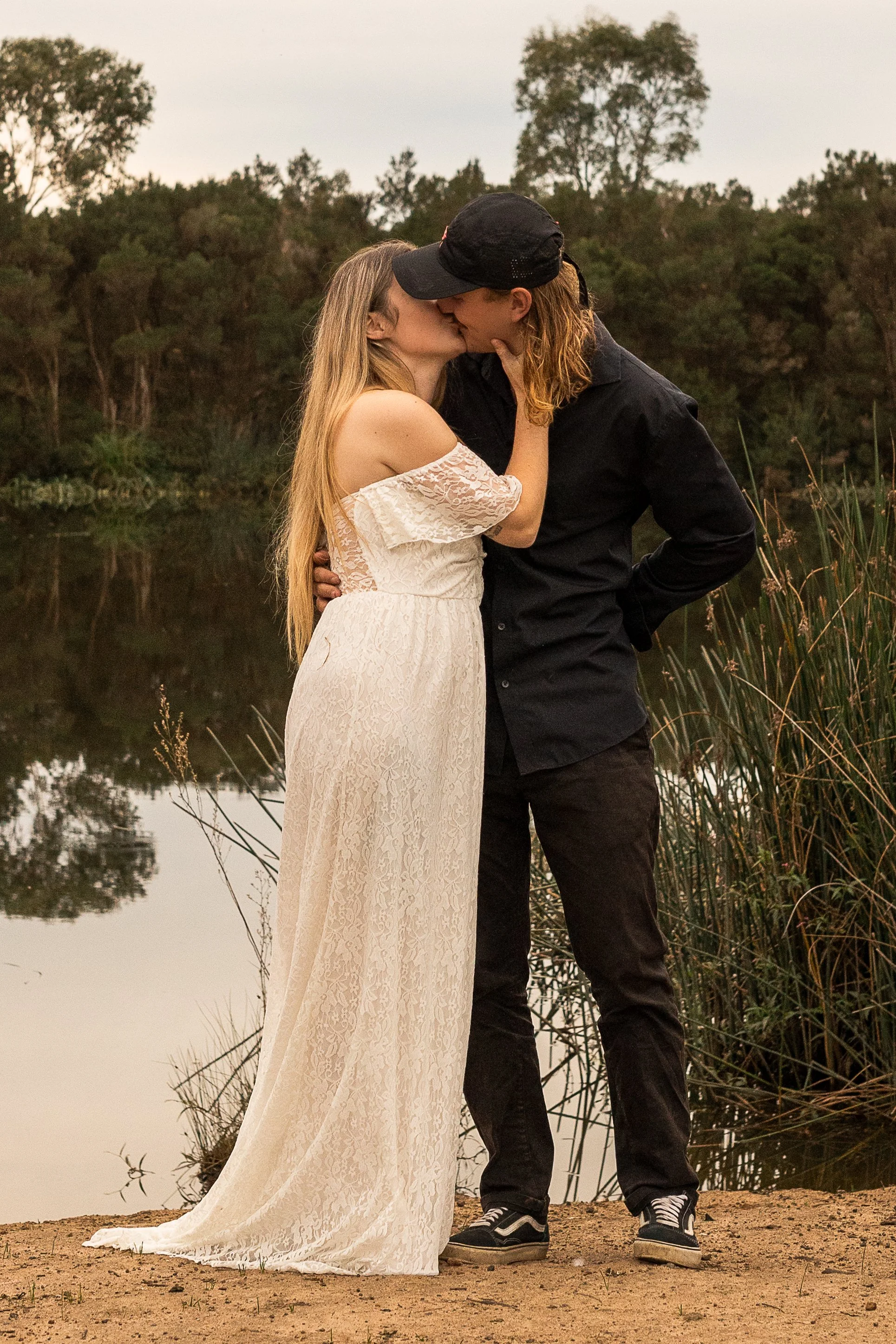 A couple kissing by a pond with trees in the background during sunset. The woman is wearing a white lace dress, and the man is dressed in black with sneakers and a baseball cap.