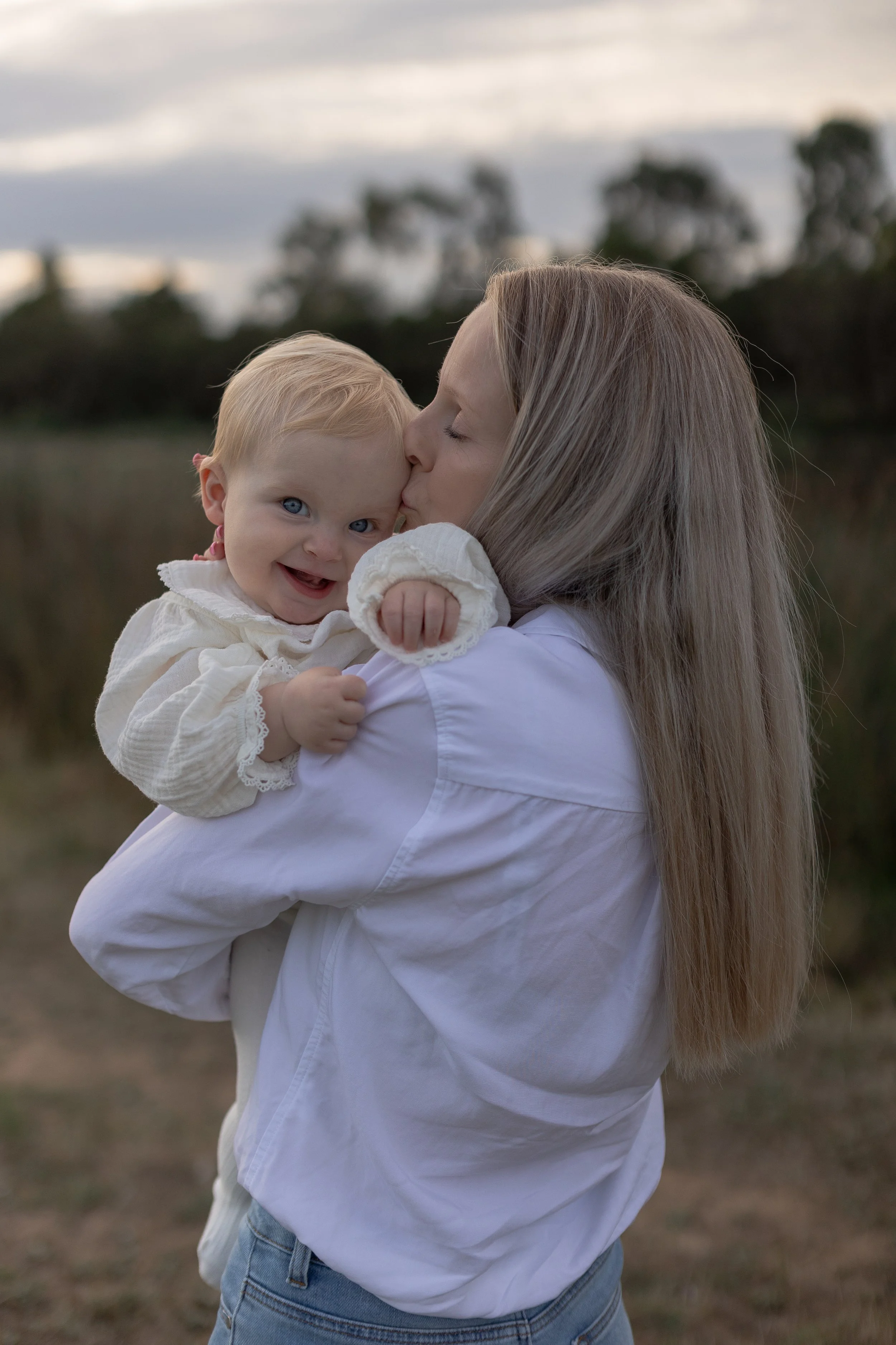 A woman with long blonde hair is holding and kissing a young girl with blonde hair, blue eyes, and a big smile, outdoors during sunset with trees and sky in the background.