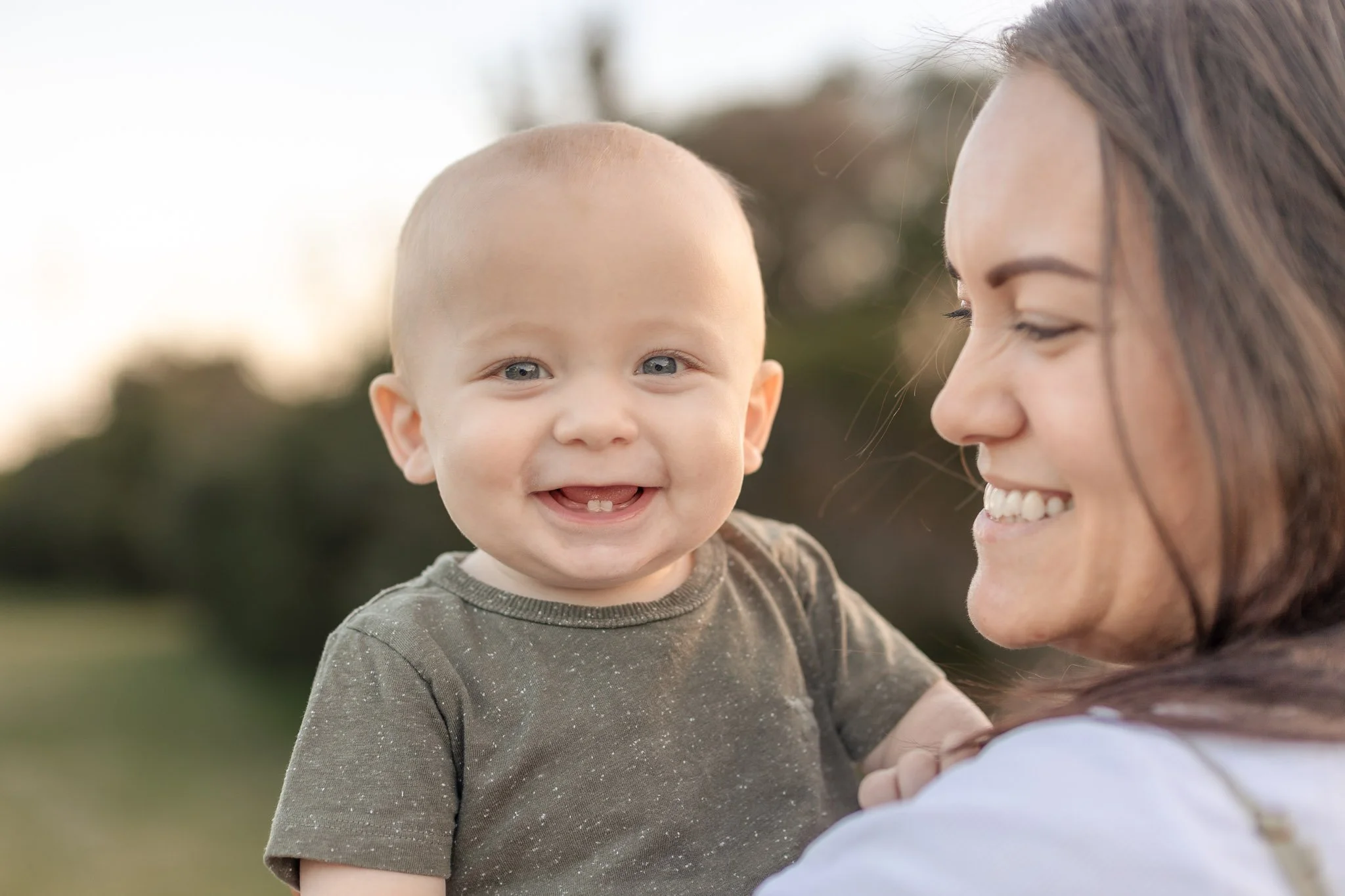 A woman smiling and holding a happy baby with blue eyes outdoors, blurred trees in the background.