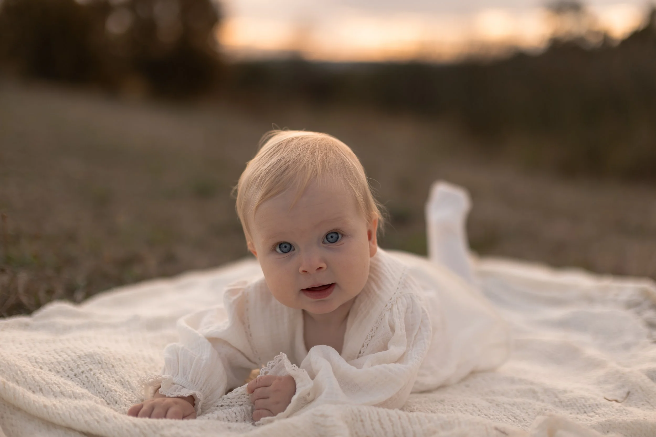 A young child with blonde hair and blue eyes lying on a blanket outdoors during sunset.