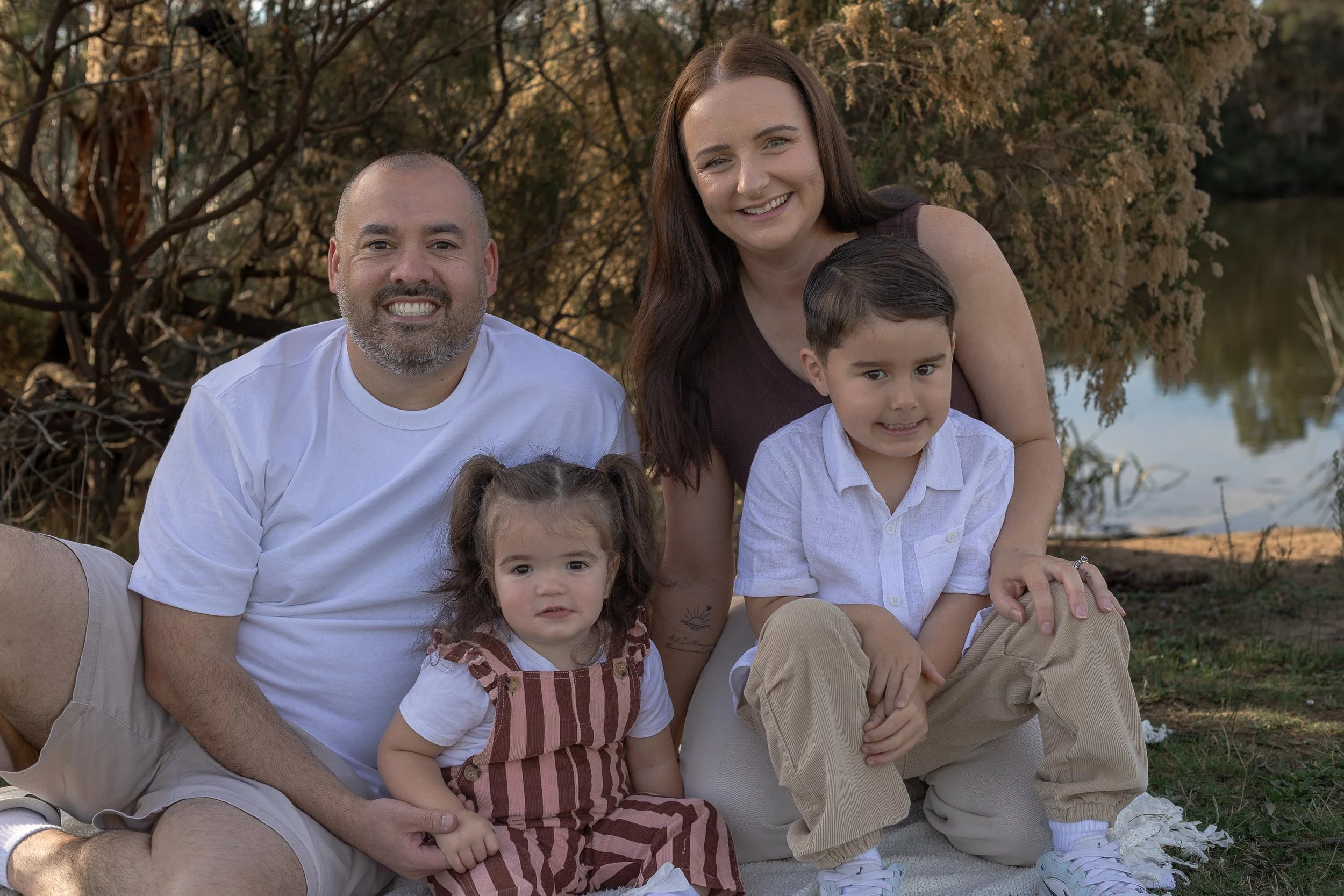 A family of five outdoor near a body of water, with trees in the background. The father is smiling and wearing a white shirt, the mother has long brown hair and is smiling, the young boy has dark hair and is in front of the mother, the young girl has brown hair styled in pigtails and is wearing a dress, and the toddler boy has dark hair, wearing a white shirt and beige pants.