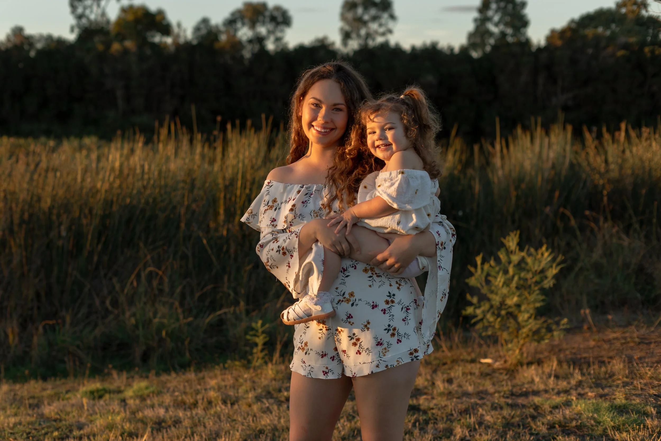 A woman with long curly hair holding a young girl with curly hair in a grassy field during sunset, both smiling warmly.