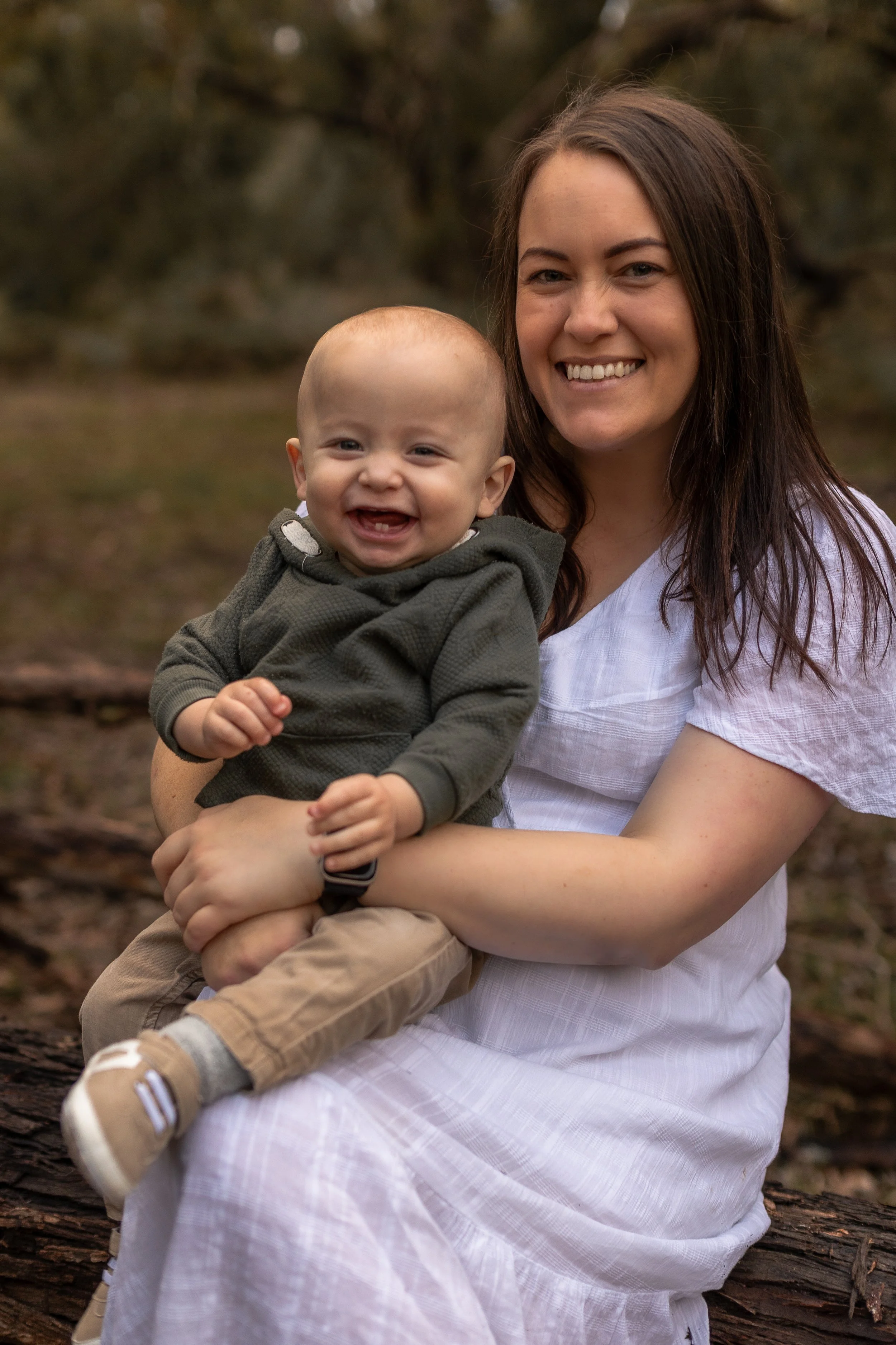 A woman holding a laughing baby outdoors with trees in the background.