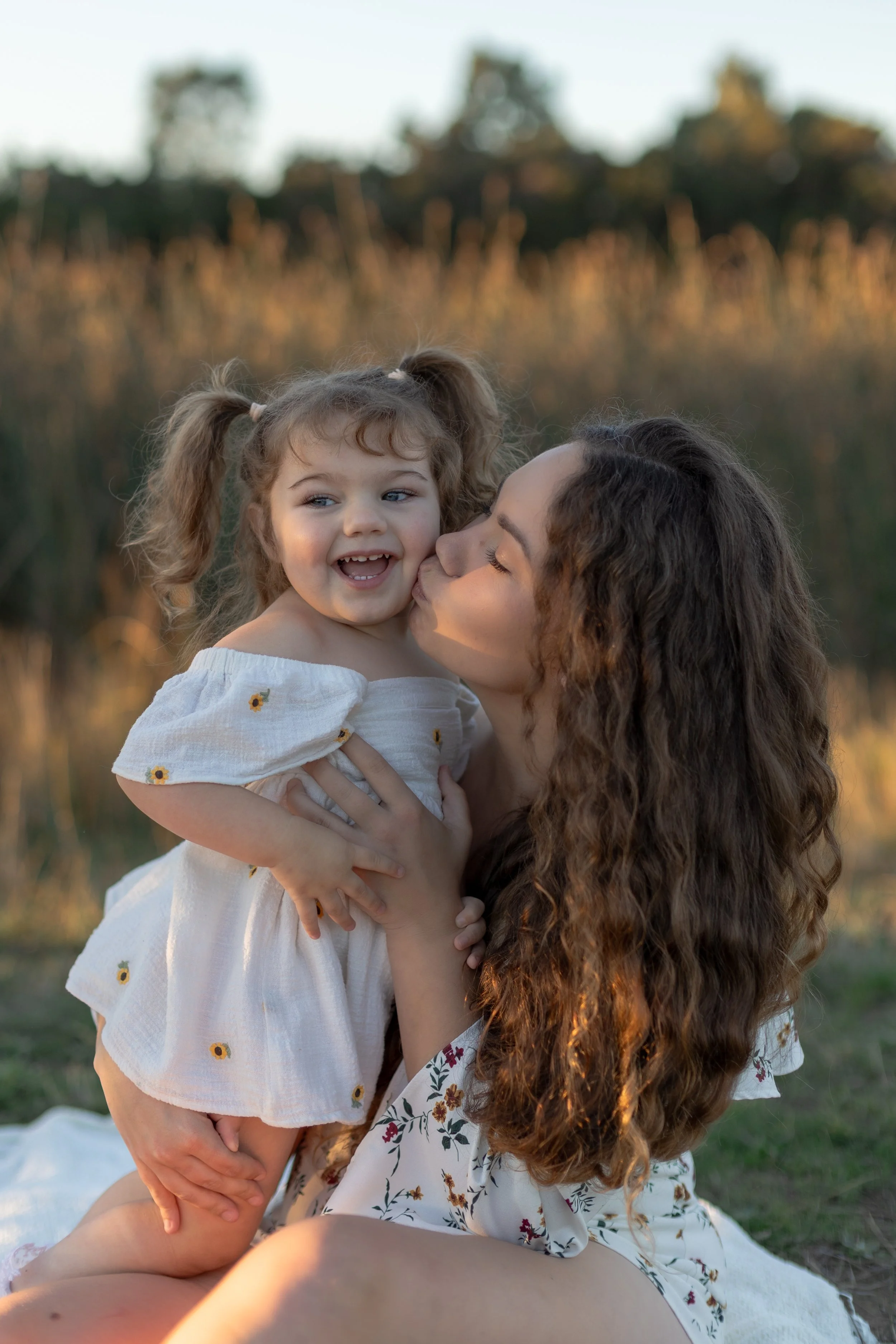 A woman with long curly hair holding a young girl with pigtails and light brown hair outdoors at sunset, both smiling and sharing a kiss.