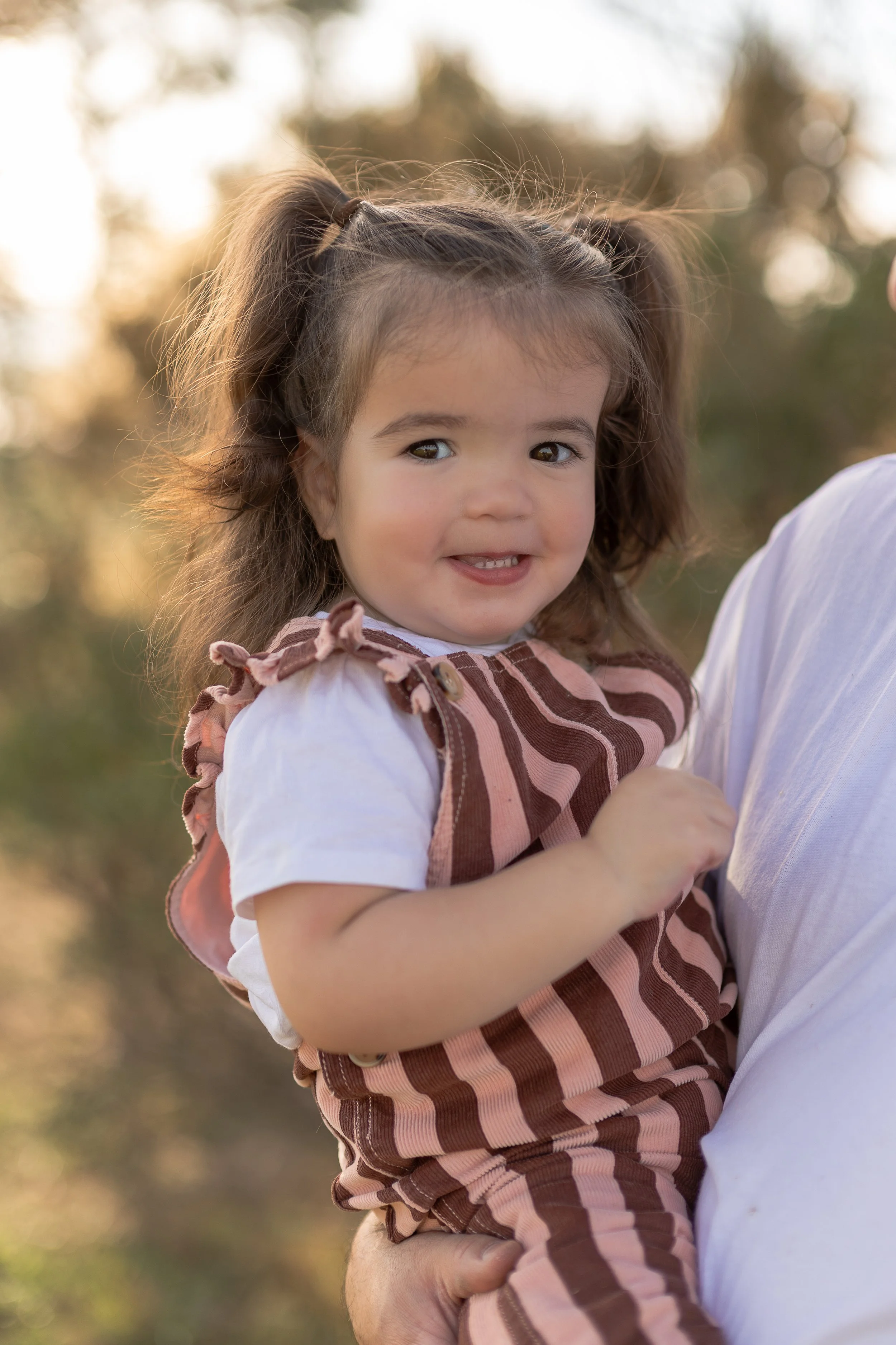 A young girl with curly brown hair tied in pigtails, smiling while being held by an adult in an outdoor setting during sunset.