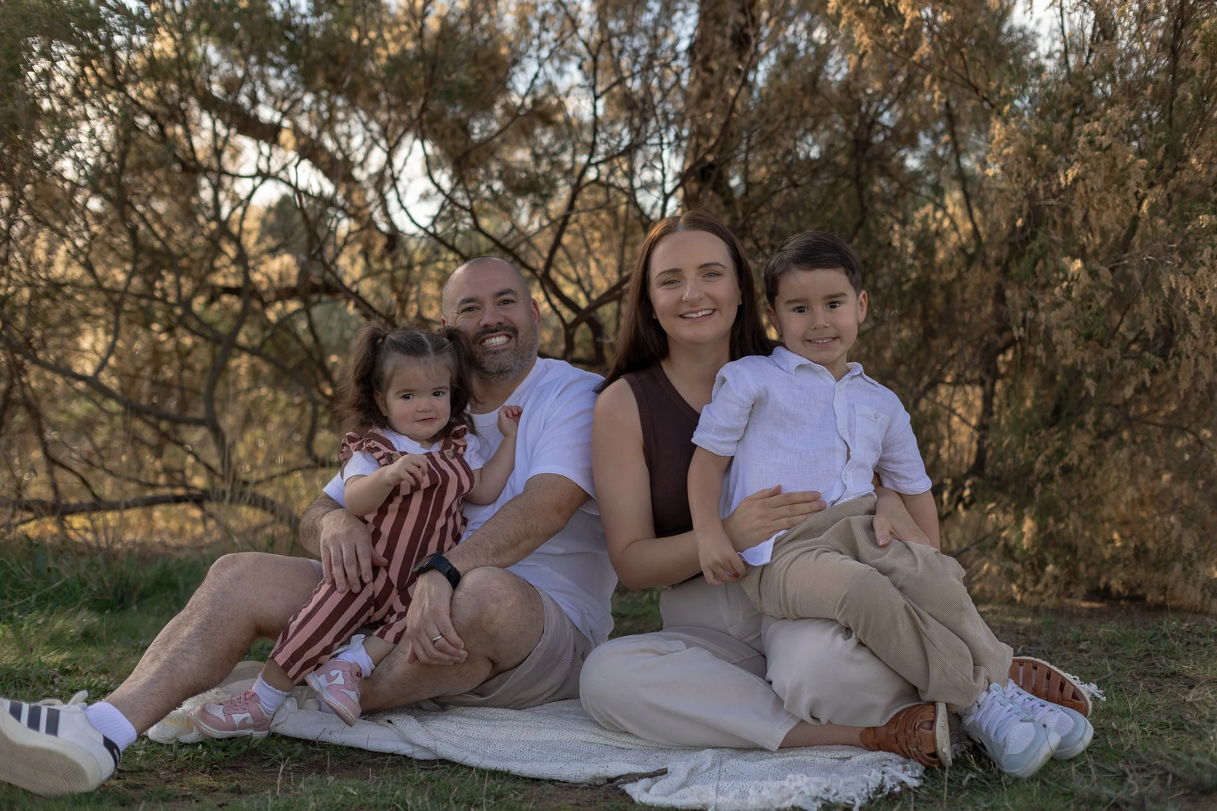 A family of four sitting on a blanket outdoors during sunset, smiling at the camera. The family includes a man, woman, a young girl, and a young boy, with trees in the background.