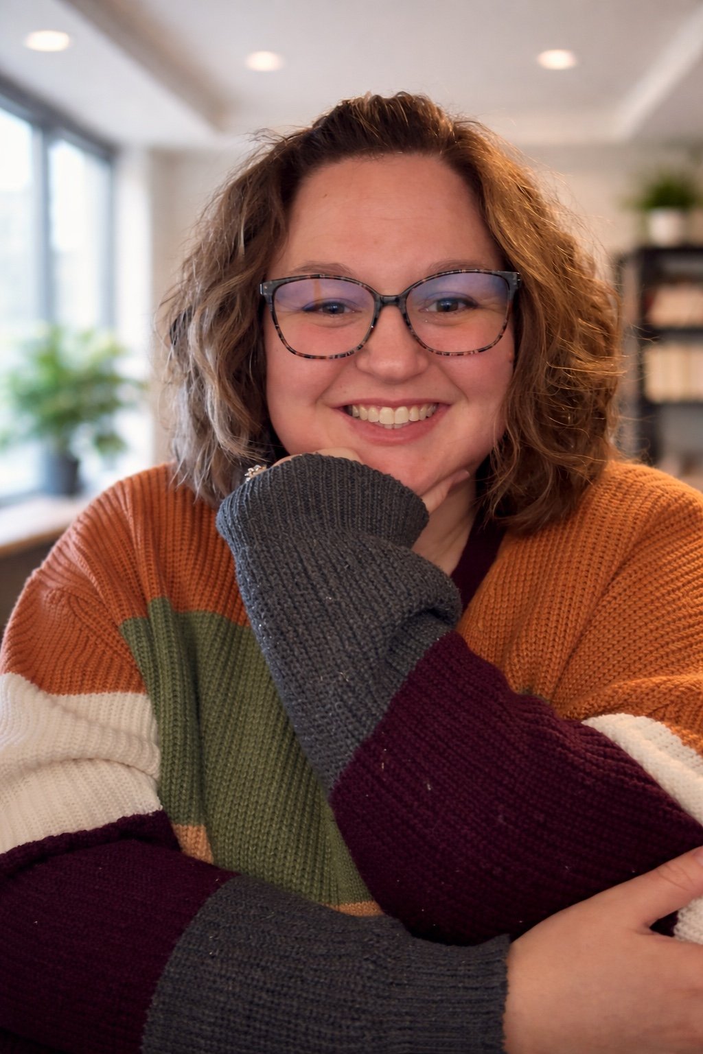 A woman with curly brown hair, glasses, and a colorful striped sweater smiling at the camera, sitting in a cozy indoor setting with natural light and plants in the background.