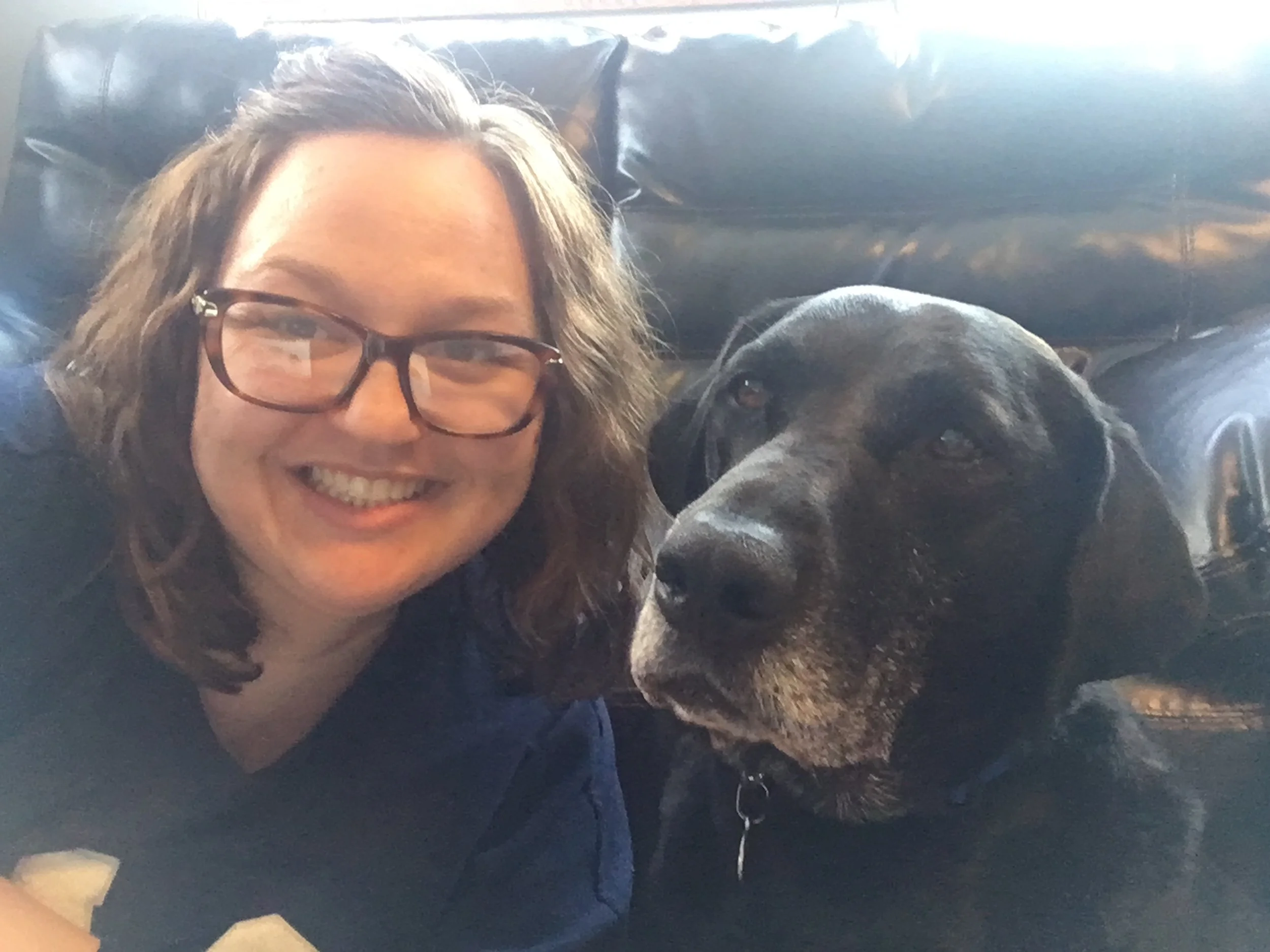 Person with glasses smiling, lying on a black pillow next to a large black dog resting on a dark surface.