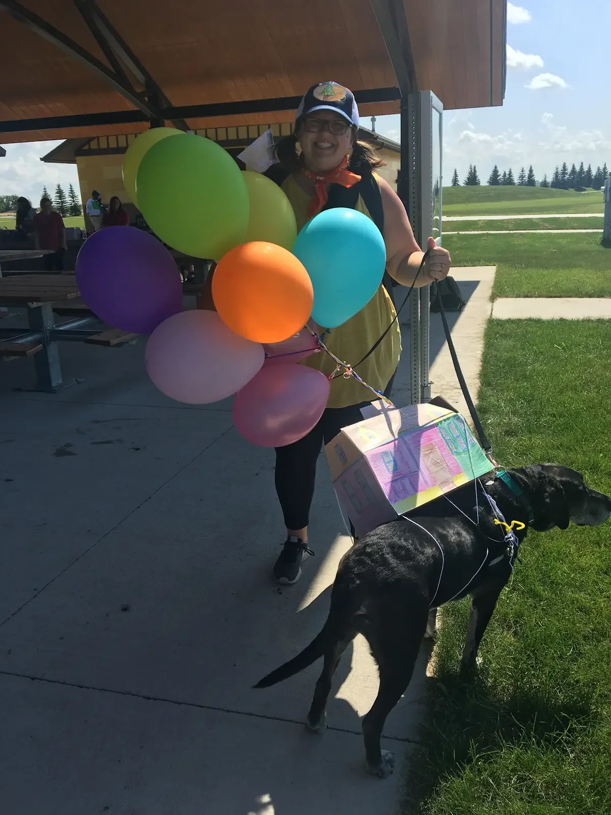 Woman holding colorful balloons and a leash attached to a black dog with a painted box on its back in an outdoor park area.