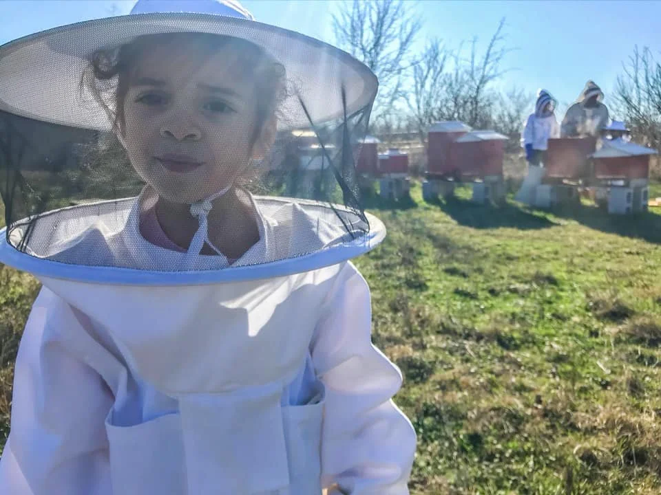 Child dressed in beekeeper suit with face net, outdoors on a sunny day, with beehives and other children in beekeeper suits in the background.