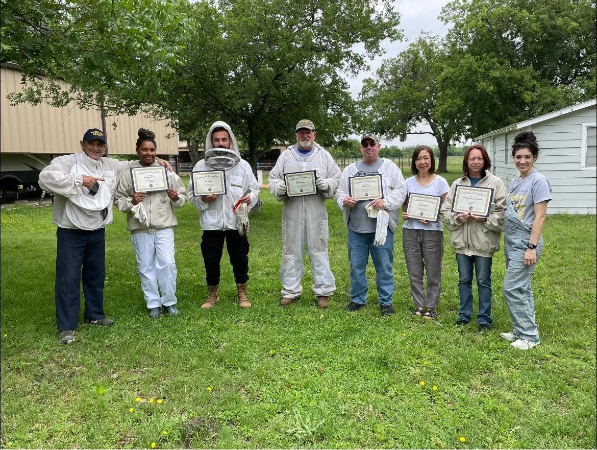 Group of eight people standing outside on grass, holding certificates, with trees and a building in the background. Two individuals are dressed in beekeeper suits, one with a hive tool and one with a smoker. Others are in casual or work clothes, smiling at the camera.